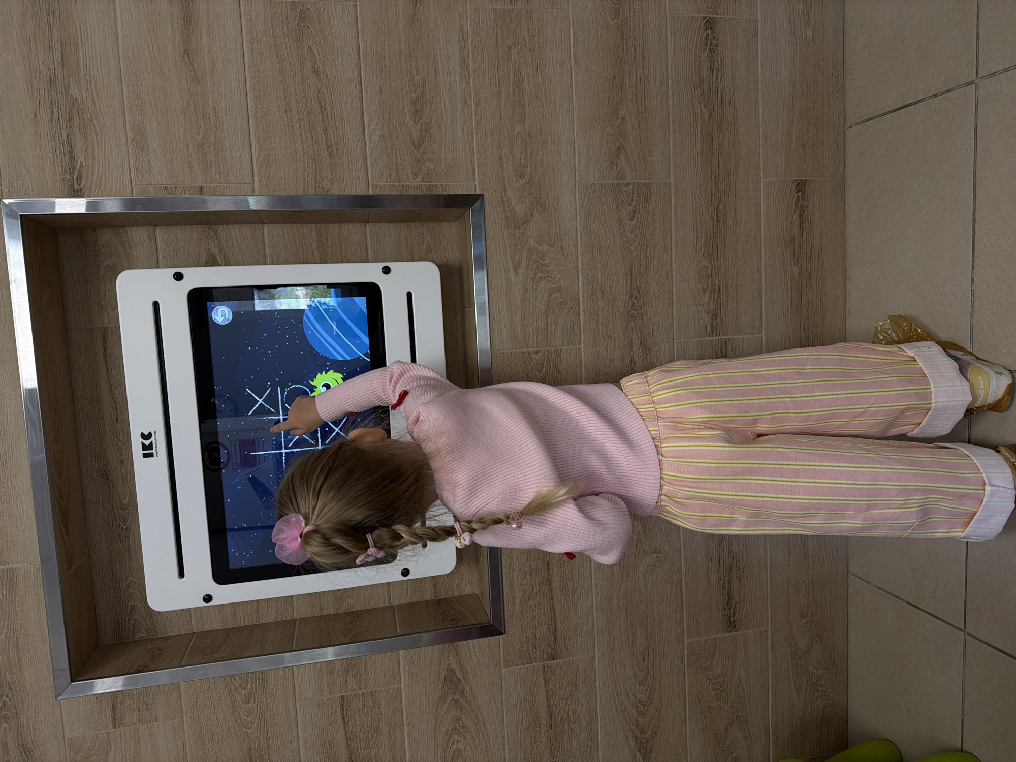 A blonde child in a pink sweater and striped pants stands in front of a floor-mounted interactive tic-tac-toe display. The display is housed within a silver-trimmed, wooden frame. The child is playing the game. The surrounding floor is tiled. The location is Kamyshin, Russia.