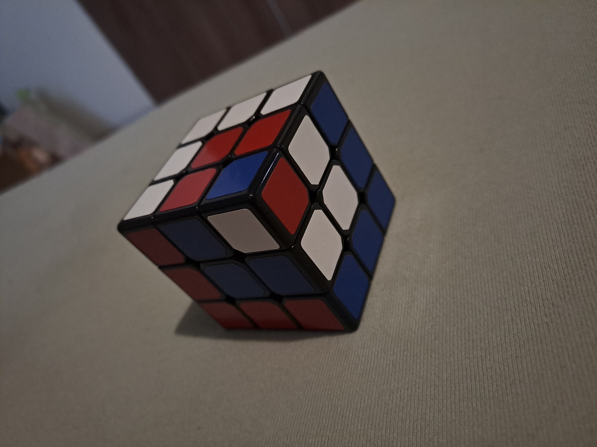 A Rubik's cube is placed on a textured light-colored surface. The cube displays white, red, and blue colored squares. The setting appears to be indoors. There is no indication of the location being Spanchevtsi, Bulgaria.
