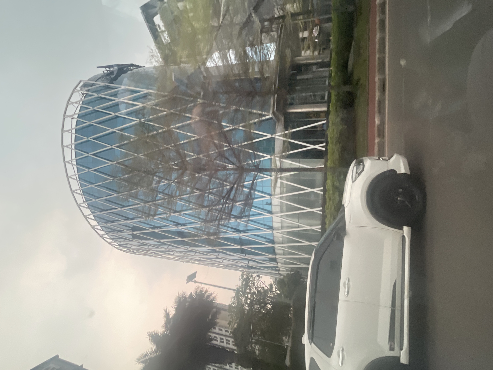 The image captures a street-level view of a modern, curvilinear building with a distinctive diamond-patterned facade in Pakulonan Barat, Indonesia. The building features large blue-tinted windows and a white structural lattice. In the foreground, the side profile of a white SUV with black wheels is visible, driving on a road. Trees, some bare-branched and others with foliage, are interspersed between the vehicle and the building. Tall apartment buildings are also present in the background to the left. The sky is overcast, suggesting a hazy or cloudy day. The scene appears to be an urban environment with contemporary architecture and vehicular traffic.