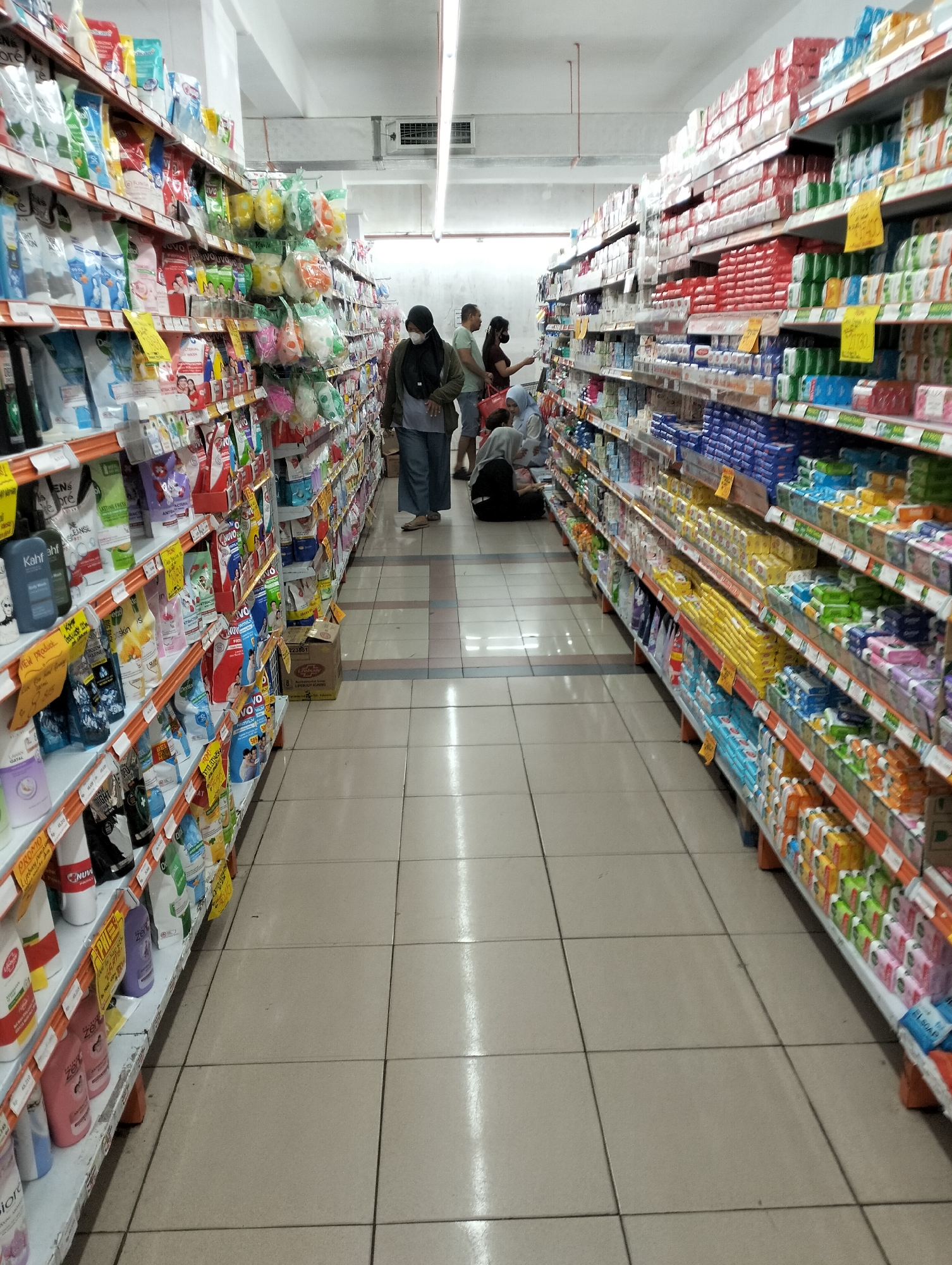 The image depicts a wide aisle in a well-stocked supermarket in Ungaran, Indonesia. The aisle is lined with tall shelves filled with a variety of personal care and household products, such as soaps, shampoos, and detergents. The shelves are organized with colorful packaging, creating a vibrant display. On the left side, a woman wearing a black hijab and a mask is walking down the aisle, while on the right, another woman is looking at the products on the shelves. In the background, a few other shoppers are visible, some interacting with the merchandise, and one person is seated on the floor. The floor is tiled, and the lighting appears to be from fluorescent overhead fixtures. The overall scene suggests a typical shopping environment in a retail establishment.