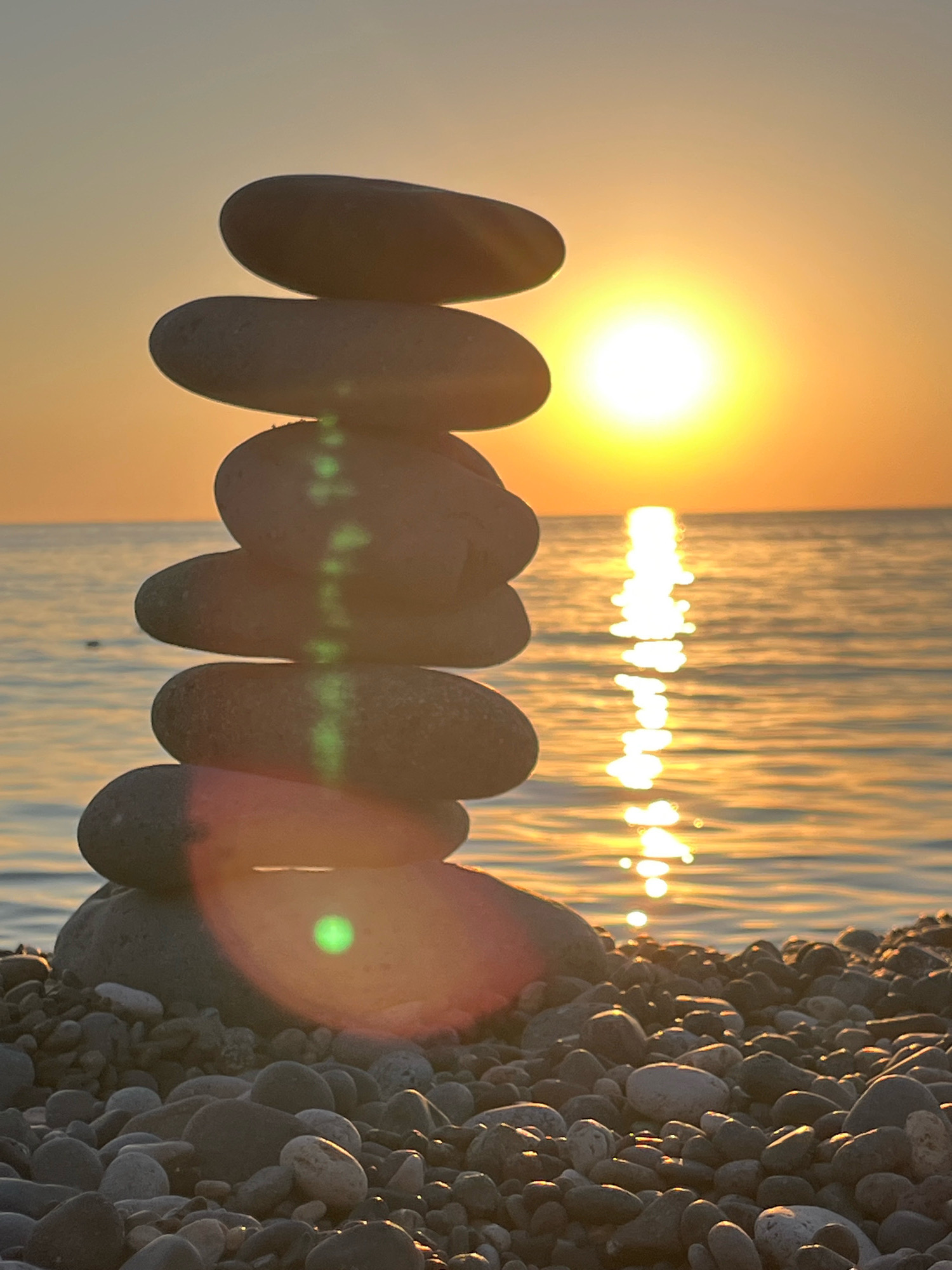 A vertical shot captures a stack of smooth, dark stones balanced on a pebble-strewn beach. The sun, a bright orb low on the horizon, casts a warm, golden glow across the scene, its reflection shimmering on the gentle waves of the Black Sea. The sky transitions from a pale yellow near the horizon to a soft, muted gray above. The stones in the stack are of varying sizes, with the largest at the base and progressively smaller ones stacked on top. A green lens flare streaks vertically through the middle of the stone stack, overlapping with some of the stones and the reflection of the sun on the water. The beach is covered in a dense layer of pebbles, some lighter in color and others dark gray, creating a textured foreground. The overall impression is one of tranquility and natural beauty, typical of a sunset scene on the coast of Kobuleti, Georgia.