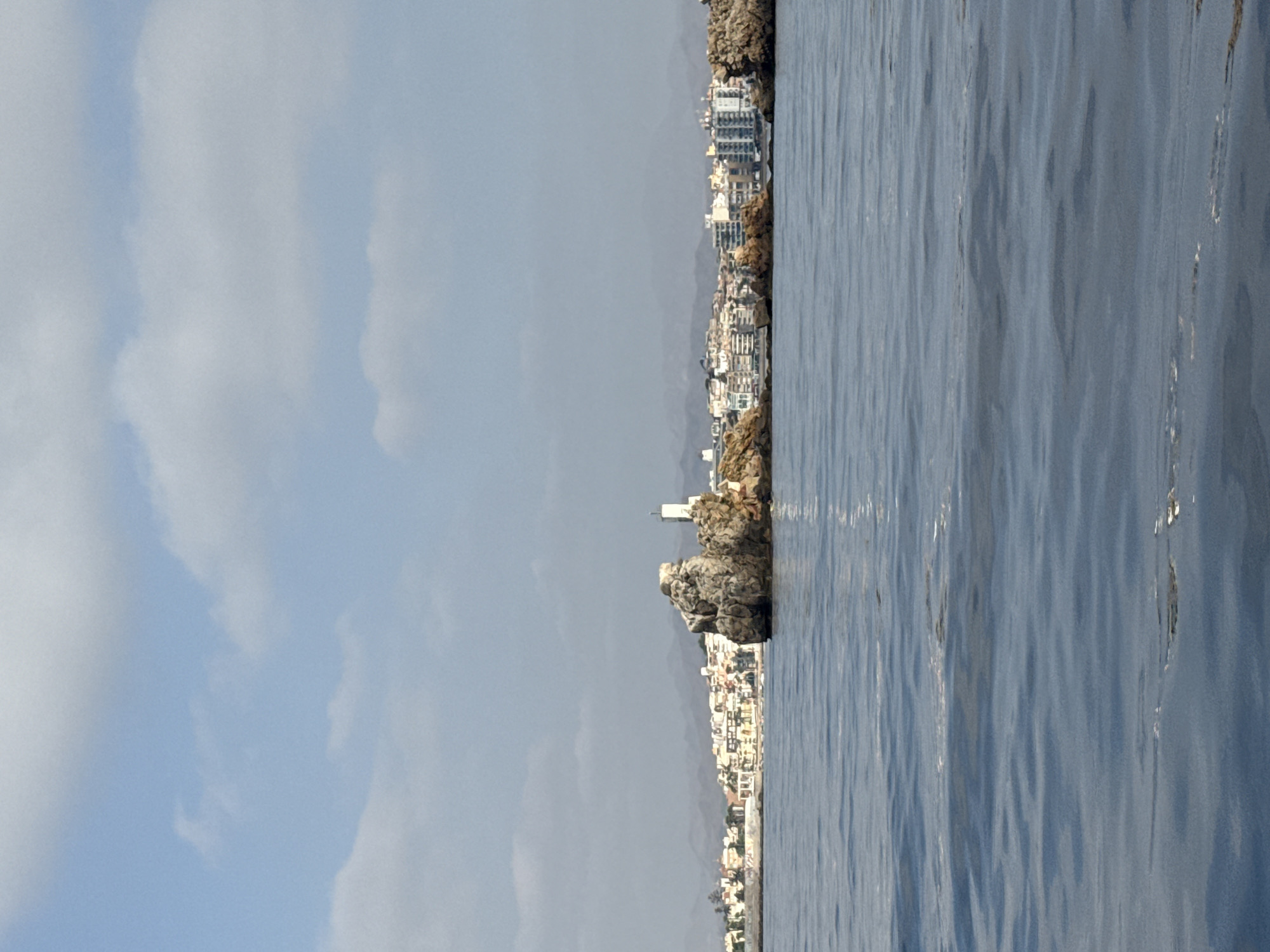 A wide-angle, eye-level shot captures a view of the Mediterranean Sea in front of Águilas, Spain. The water, a deep blue, is calm with subtle ripples. In the middle ground, a cluster of rocky outcrops jut out from the sea, one of which features a small, white lighthouse. Behind the rocks, the coastline of Águilas is visible, a strip of white and pastel-colored buildings with a backdrop of hazy, mountainous terrain. The sky above is a pale blue, dotted with scattered white clouds. The image conveys a sense of tranquility and the coastal beauty of the region.