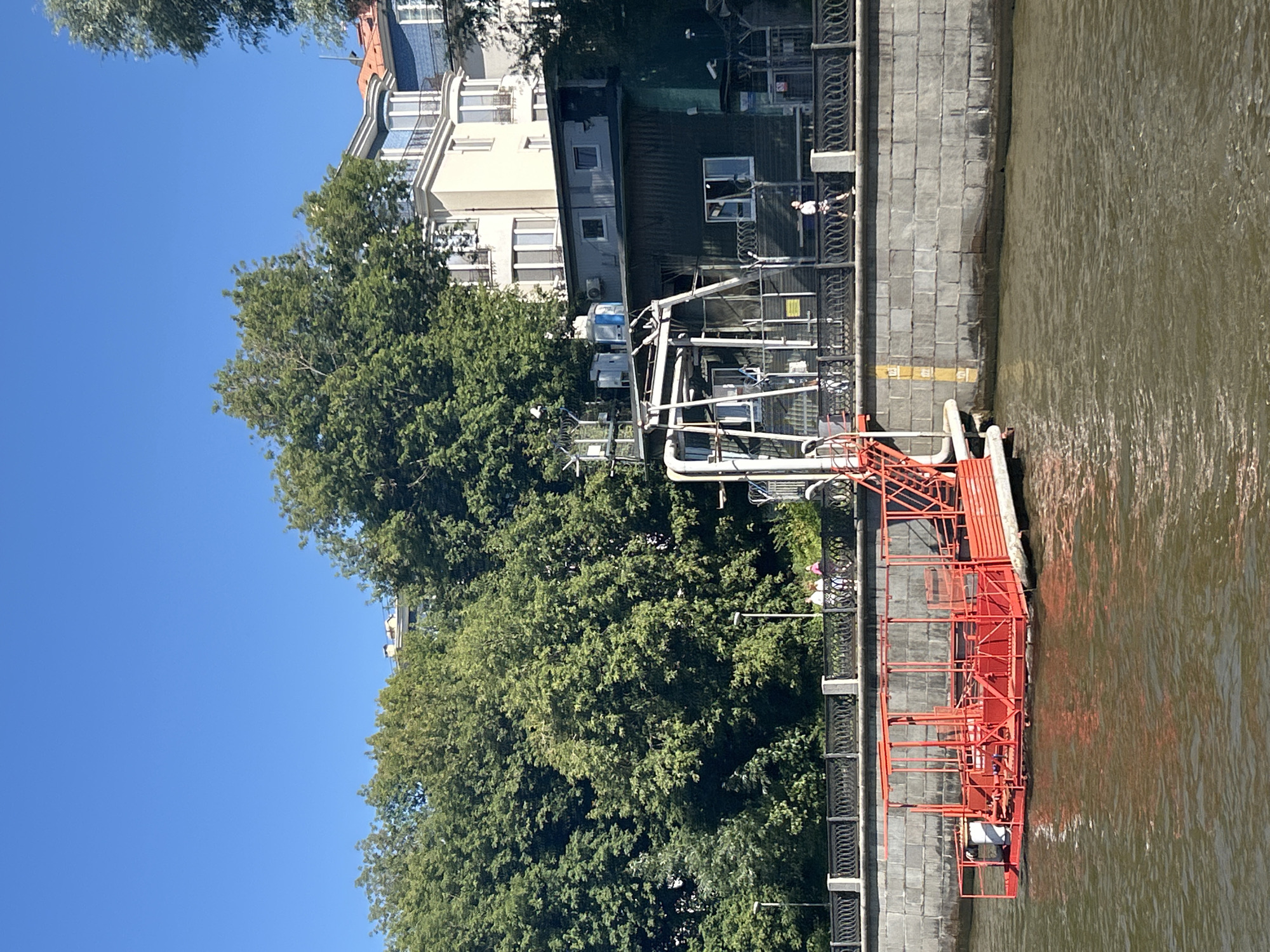 This image shows a view of the Moscow River on a clear day. In the foreground, a bright red platform or boat is docked. The platform has a cage-like structure with railings and stairs leading up to it. The water is a murky brown. Behind the platform and riverbank, there is a stone embankment with a decorative black metal fence. Beyond the fence, people are walking along a pathway. Several trees with lush green foliage are visible, and behind the trees, there are buildings, including a light-colored building with balconies and a darker, corrugated building with windows. The sky is a vibrant, clear blue. The scene appears to be in a well-developed urban area.