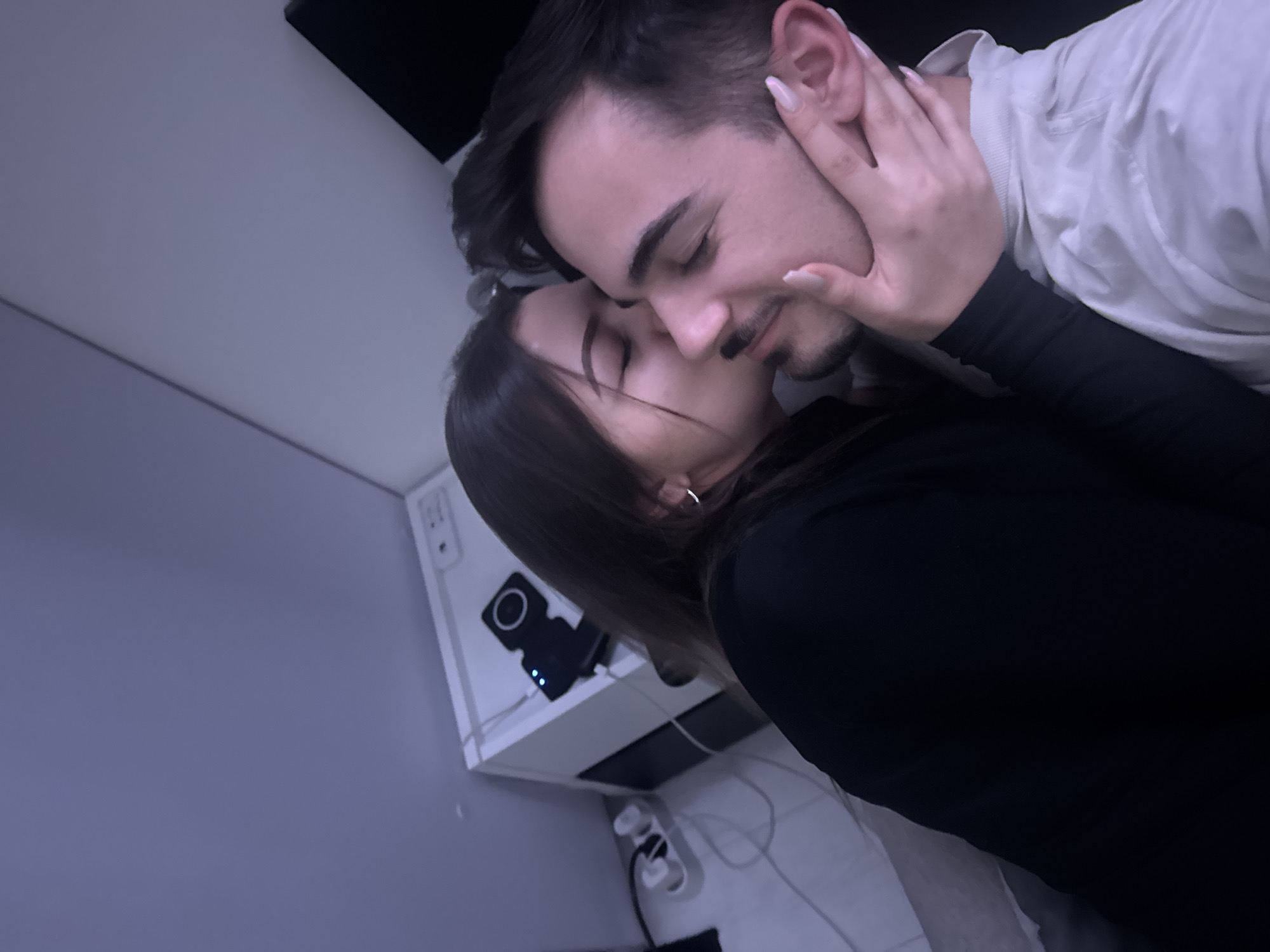 A close-up, dimly lit indoor photograph features two individuals in an intimate pose. A female, with long dark hair and wearing a dark long-sleeved top, is positioned on the left, her eyes closed, kissing the male subject on the right side of his head/face. Her left hand, adorned with light-colored manicured nails, gently cups the male's jaw and neck. She wears a small hoop earring on her visible ear. The male subject, with dark hair, a mustache, and a short beard, is wearing a light-colored (possibly white or light grey) collared garment, with his eyes closed.
The immediate background to the left of the female shows a white, cube-shaped piece of furniture, possibly a nightstand. On its surface, a black electronic device with a circular design and three illuminated blue indicator lights is present. Below this furniture, a white power strip with multiple plugs and associated wiring is visible on the floor. To the upper right, behind the male's head, a dark, rectangular object is partially obscured. The walls are a neutral light grey or bluish-grey. The specific location within the city of Sofia, Bulgaria, cannot be determined from the visual information provided in the image.