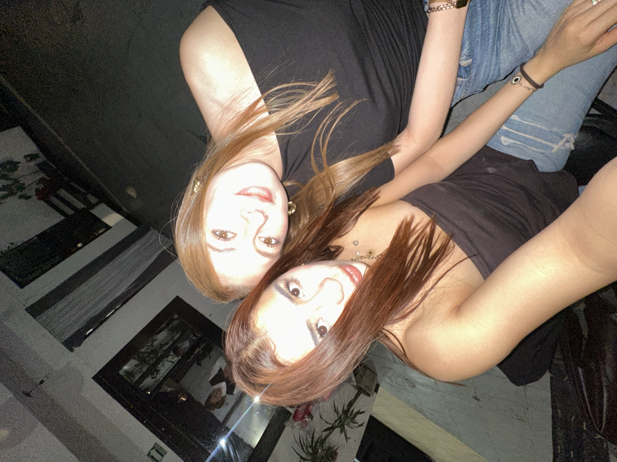 Two women are in the image. One woman has reddish-brown hair and is wearing a black top. The other woman has light brown hair and is also wearing a black top and jeans. They are both looking at the camera. The background appears to be an outdoor setting, possibly in San Bernardino Tlaxcalancingo, Mexico, with architecture and plants visible.