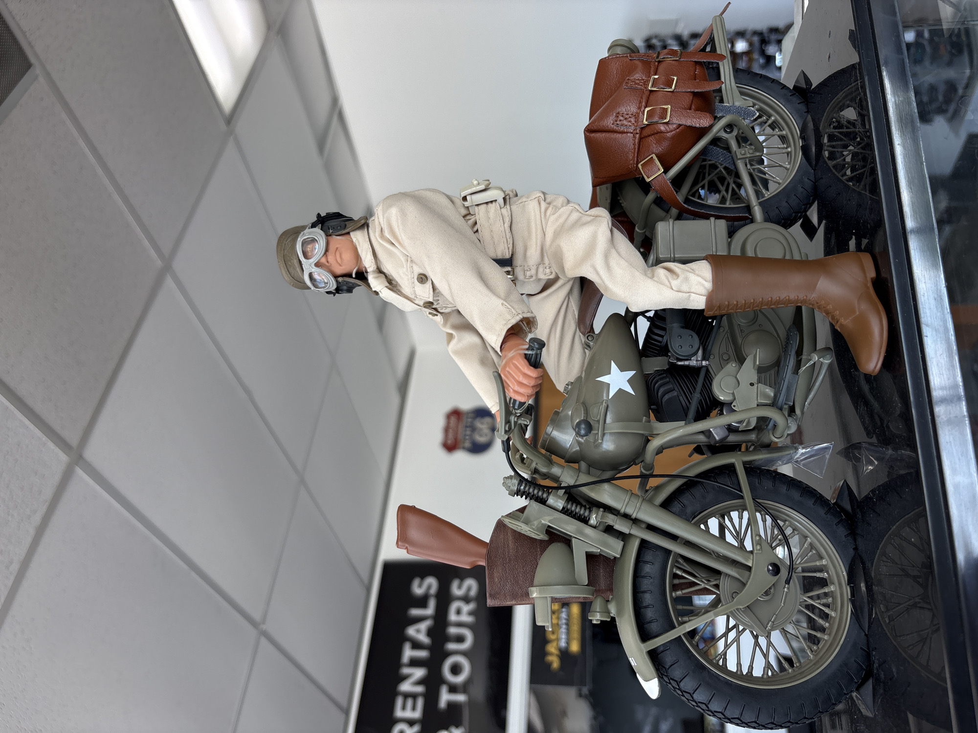 A realistic action figure of a man in vintage military attire, complete with a helmet, goggles, and a tan uniform, is posed riding a detailed replica of a vintage olive-drab motorcycle. The motorcycle is equipped with leather saddlebags and a rifle case mounted on the front. The action figure and motorcycle are displayed on a reflective black surface, possibly a display case, in what appears to be an indoor commercial space. In the background, signage can be seen, including a black sign with white text reading "RENTALS & TOURS" and a smaller yellow sign with "JACKE RENTAL." A Route 66 sign is also visible. The ceiling features suspended acoustic tiles and fluorescent lighting, typical of a retail or office environment. The scene is illuminated by overhead lighting, casting a bright light on the subjects and creating reflections. The overall impression is a curated display of a historical military scene. The location is identified as Des Plaines, United States of America.