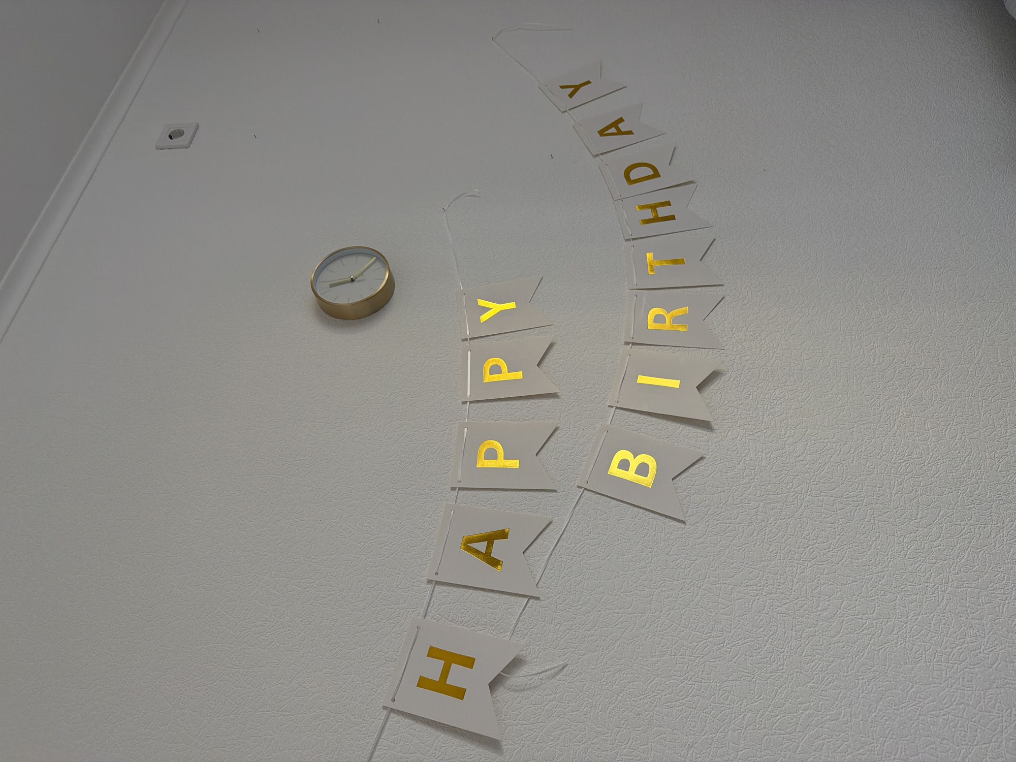 A gold analog clock is mounted on a textured white wall, slightly to the right of the frame's center. Below the clock, two strings of white pennant banners with gold lettering are strung across the wall. The upper banner reads "HAPPY" from left to right, with the letters "H", "A", "P", "P", and "Y" clearly visible. The lower banner, positioned below and to the right of the upper banner, reads "BIRTHDAY" from left to right. The visible letters on the lower banner are "B", "I", "R", "T", "H", "D", "A", and "Y". A white electrical outlet is visible on the wall to the upper right of the clock. The overall environment appears to be indoors, likely a room in a residential or public building. There is no discernible location in the city of Kyiv, Ukraine specified in the image.
