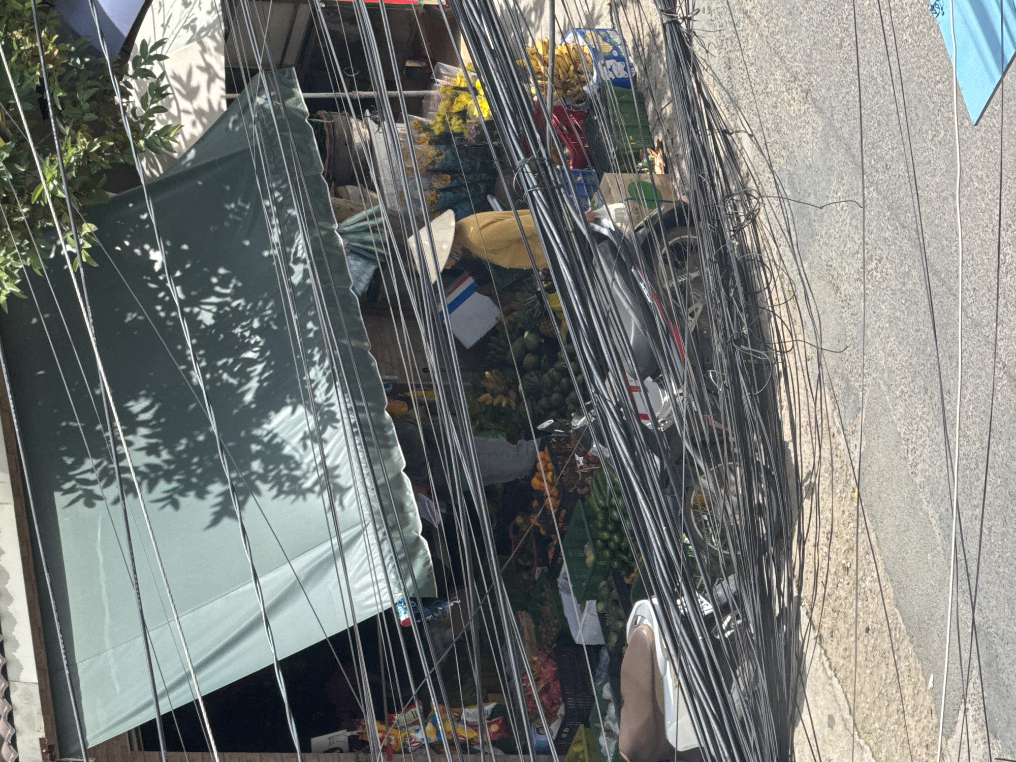 This image captures a bustling street scene in Da Lat, Vietnam, likely a market area characterized by a dense network of overhead electrical wires. The foreground is dominated by a thick tangle of these wires, obscuring much of the view. Behind them, a market stall is visible, featuring a variety of fruits displayed in crates, including bananas, dragon fruit, and oranges. A person wearing a traditional conical hat and a yellow shirt is standing at the stall, interacting with the produce. A motorcycle is parked nearby, also partially hidden by the wires. The market stall is covered by a green awning, and the dappled sunlight filtering through the leaves of an unseen tree creates shadows on the awning and the ground. The overall impression is one of vibrant activity within a densely populated urban environment.