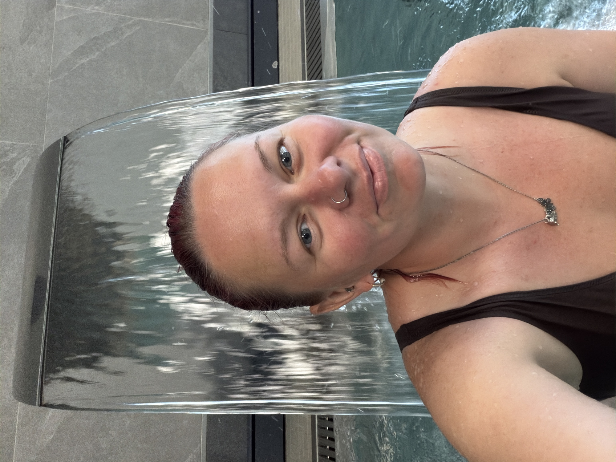 A woman is submerged in a pool of water, likely a spa or similar establishment, with a cascade of water flowing over her. The woman, who has fair skin and short reddish-brown hair, is wearing a dark-colored swimsuit and a delicate necklace. She has a nose ring and is looking directly at the camera with a slight smile. The water is clear and reflects the surrounding environment. The pool appears to be constructed with gray tiled walls and a dark metal finish on the waterfall feature. The location is identified as Chaikovsky, Russia, based on external information.
