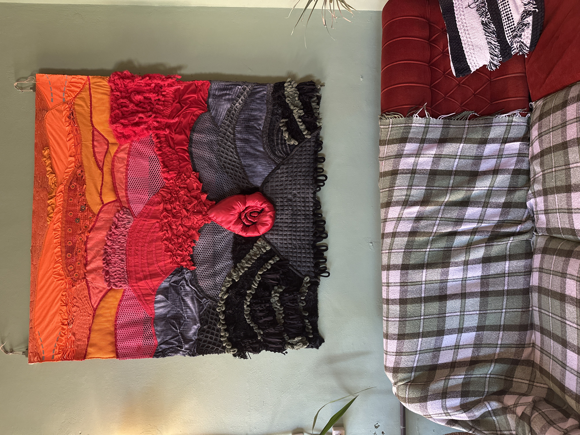 The image depicts a wall with a textured fabric artwork hanging on it. The artwork is rectangular and features a gradient of colors, from orange to red to gray and black. The textures vary from smooth to ruffled to mesh-like. A red fabric rose is attached to the gray and black section. To the right of the artwork is a red tufted sofa, partially covered with a green and white plaid blanket. The wall is light green. There is no evidence to suggest that this image was taken in Kryvyi Rih, Ukraine.