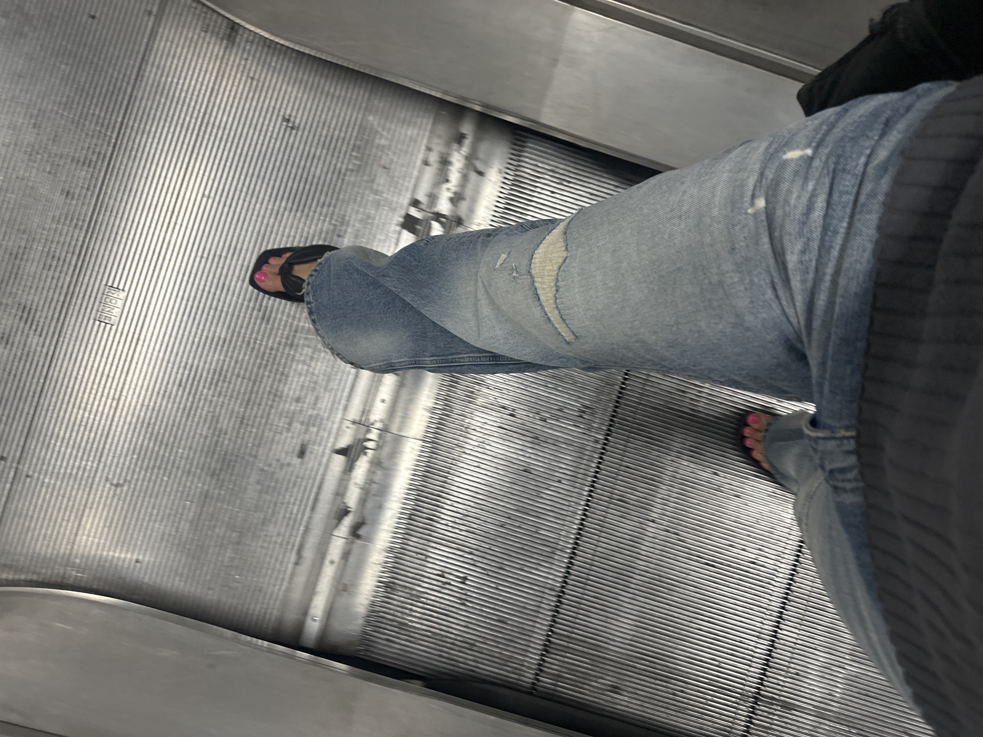 The image shows a person wearing blue ripped jeans and black flip-flops on an escalator. The person's toes are painted pink. The escalator has a metal grooved surface and appears to be in a public space. The precise location within London, United Kingdom, cannot be determined from the image.