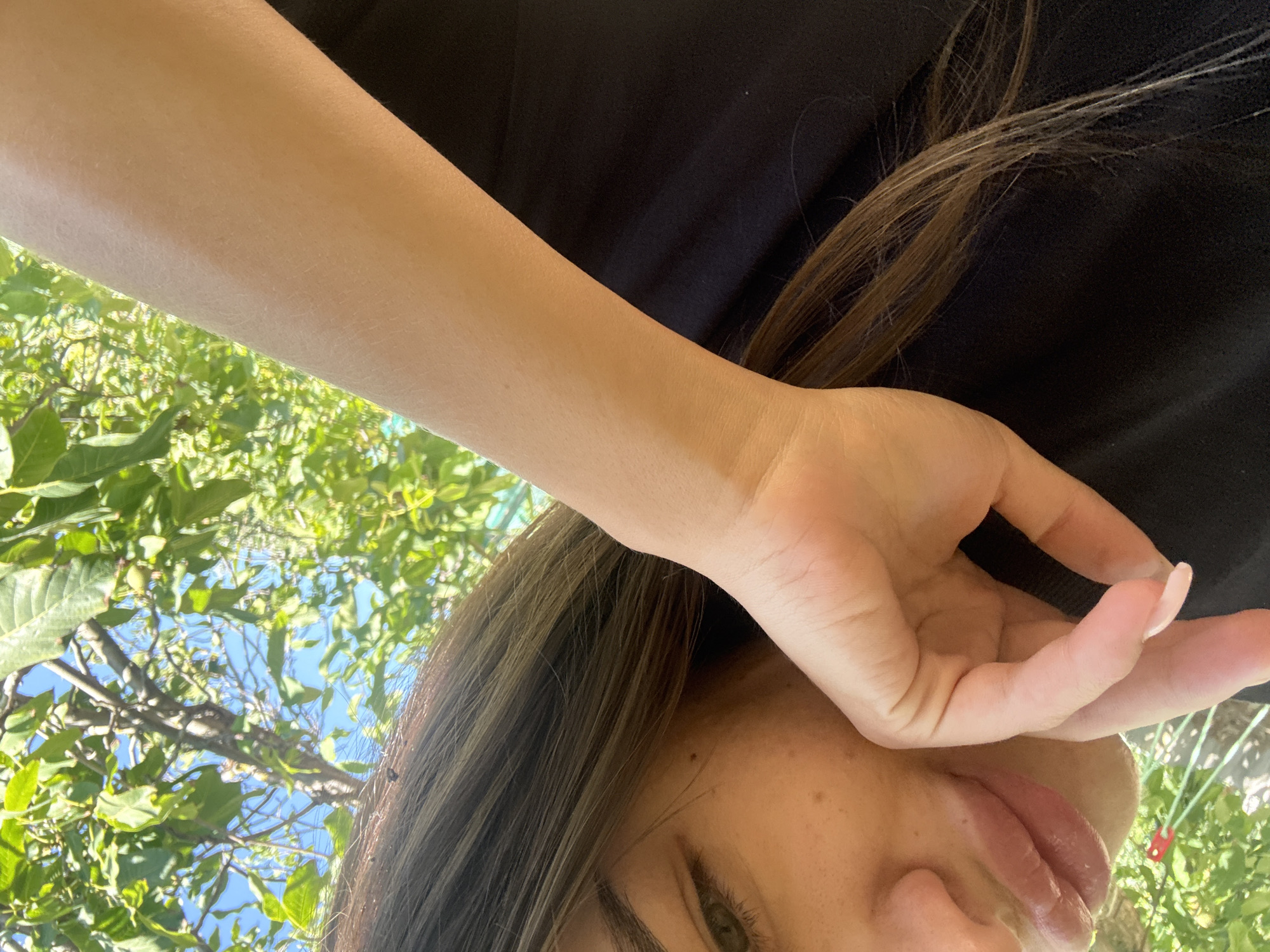 The image is a close-up, from a low angle, of a person's face and arm against a backdrop of green leaves and blue sky. The person, partially visible, has fair skin and light brown hair. Their lips are glossy and slightly parted. One hand is raised, with the fingers gently touching the cheek or chin, and a light-colored nail polish is visible on one nail. The arm extends diagonally across the frame, its skin catching the sunlight. The background consists of lush green foliage and a clear blue sky, suggesting an outdoor setting, possibly in a park or garden. There is no indication of the city of Basarabeasca, Moldova, within the image itself.