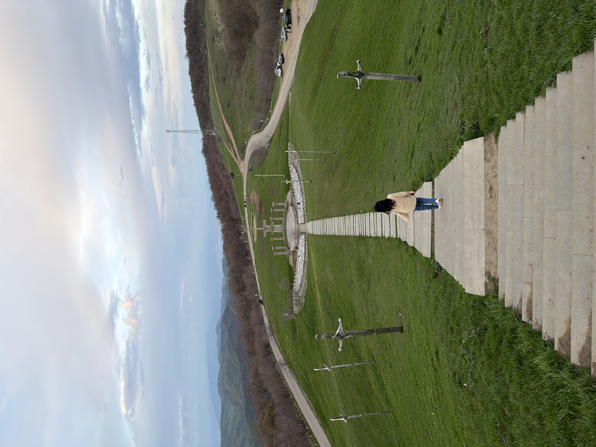 A wide, angled shot shows a woman walking down a concrete staircase in the Memorial of Georgian Warriors near Zemo Nichbisi, Georgia. The woman wears a light-colored coat, jeans, and light-colored shoes. She has dark hair and is seen from the back. The staircase descends through a grassy area dotted with sword-shaped monuments made of wood and metal. To the left of the stairs, there is a walking path, trees and the sky. In the distance, there are hills, a tower, and several parked cars.