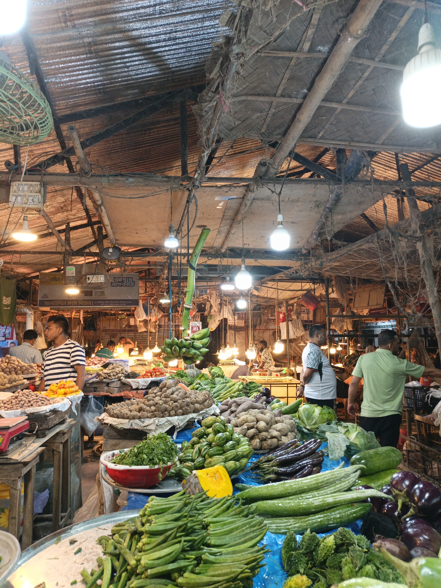 The image depicts a bustling marketplace in Dhaka, Bangladesh, likely a local bazaar. The location is suggested by the environment. The market is housed under a partially dilapidated corrugated metal and bamboo roof structure with exposed wiring, light bulbs and fans.
Various vendors are displaying produce including potatoes, tomatoes, okra, gourds, eggplants, cabbage, and bananas. Customers browse and interact with vendors. One man wears a black and white striped shirt, while another wears a green shirt. The scene is filled with a variety of goods for sale.
