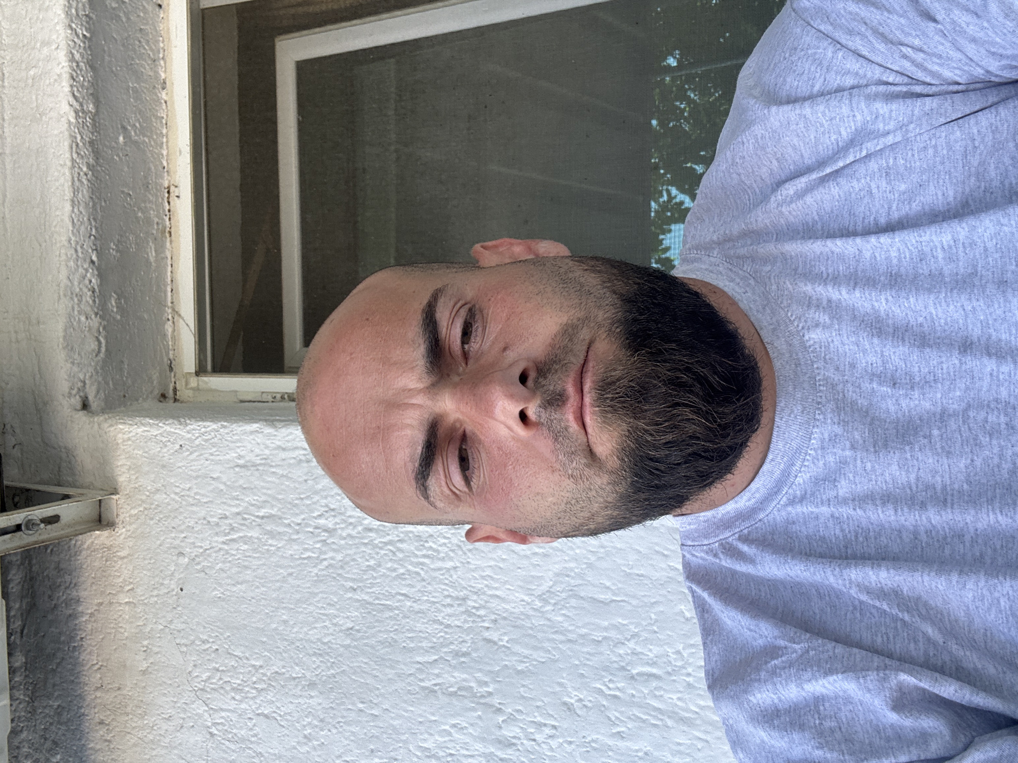 A medium shot depicts a man with a bald head and a full, dark beard. He is wearing a light gray long-sleeved shirt. His expression appears serious or thoughtful. The background includes a white, textured wall, part of a window frame, and a glimpse of foliage. There is no indication of a location in the city of Sarnevo, Bulgaria, as nothing in the image suggests a specific location.