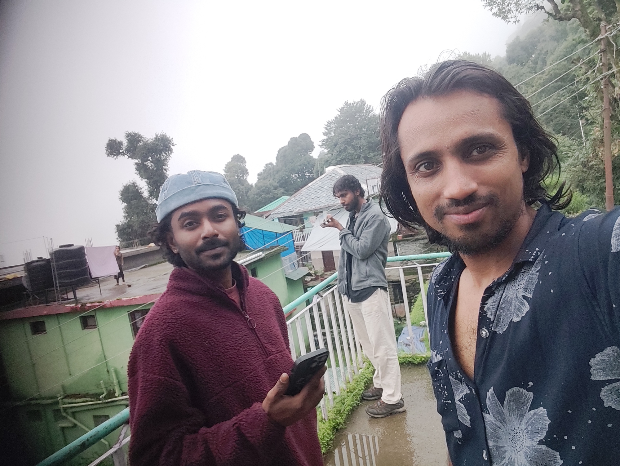A medium shot captures three men on a rooftop in Haini, India, on a cloudy day. The man in the foreground on the right, closest to the camera, is wearing a dark blue shirt with a floral pattern and has long, dark hair. He is looking directly at the camera. To his left, a man in a light blue beanie and a maroon fuzzy zip-up jacket is holding a smartphone in his right hand and looking down at it. Behind them, a third man in a light gray jacket and off-white pants is standing with his hands clasped in front of him and looking away from the camera. The background shows a multi-story building with a green facade, a tiled roof, and water tanks on the roof. Trees and mountains are visible in the distance, suggesting a hilly or mountainous location. The ground appears wet, indicating recent rain.