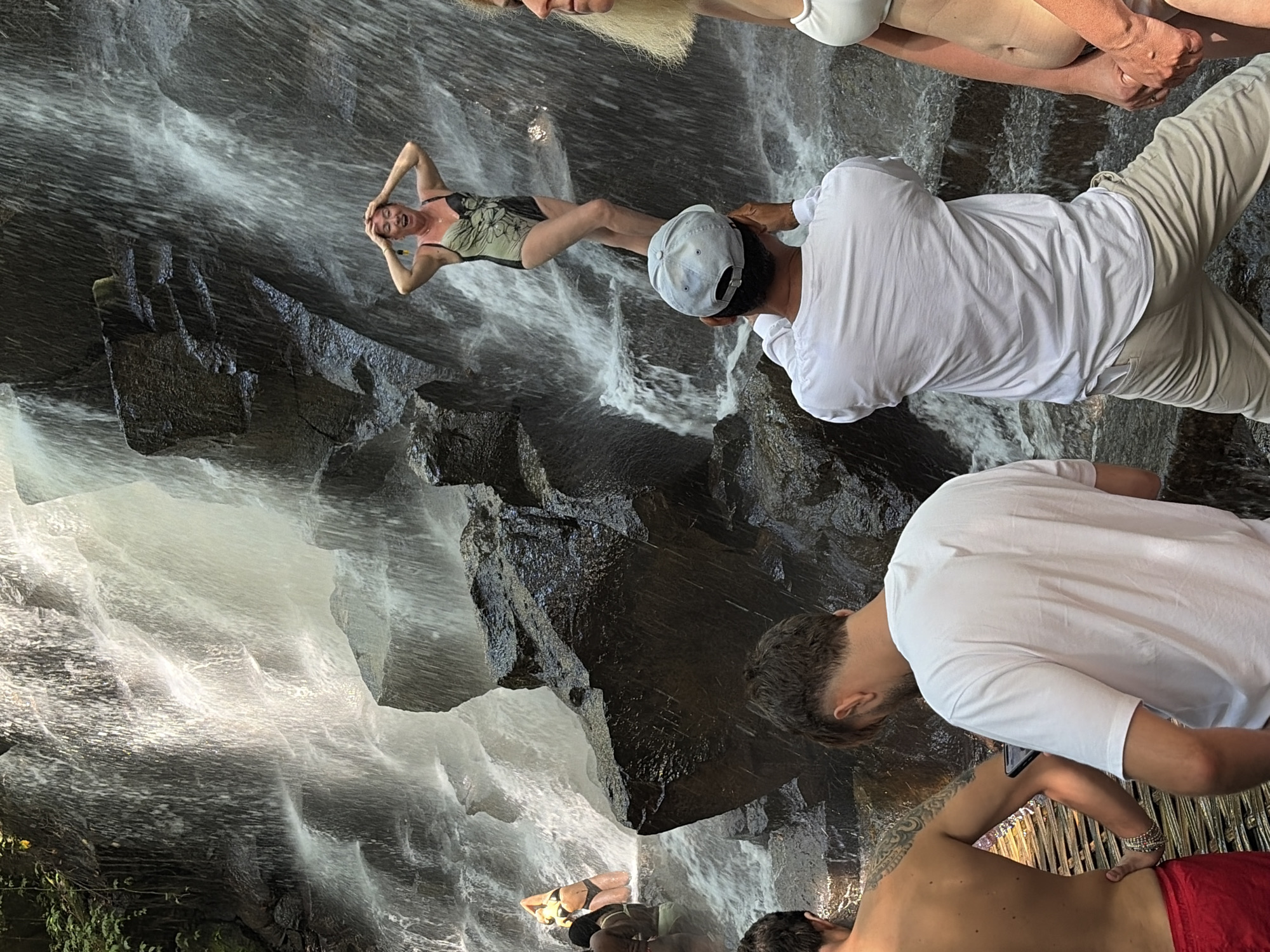In the city of Gianyar, Indonesia, a group of people are gathered at the base of a cascading waterfall. The waterfall flows over dark, wet rocks, creating a misty and dynamic environment.
In the foreground, two men are partially visible from the back. One, on the left, has tattoos on his back and is wearing red shorts. He is holding his left hand on his hip. The other man, in the center, is wearing a white long-sleeved shirt and beige pants, and he is looking down at a mobile device he is holding.
Behind them, a woman in a one-piece swimsuit with a floral pattern is standing on a rock in the middle of the waterfall, with her hands on her head and her mouth open. To her right, another woman, with blonde hair and a white bikini top, is standing and holding hands with an unseen person. Further to the left, another woman in a bikini is posing near the water. The overall scene suggests a recreational visit to a natural attraction.
