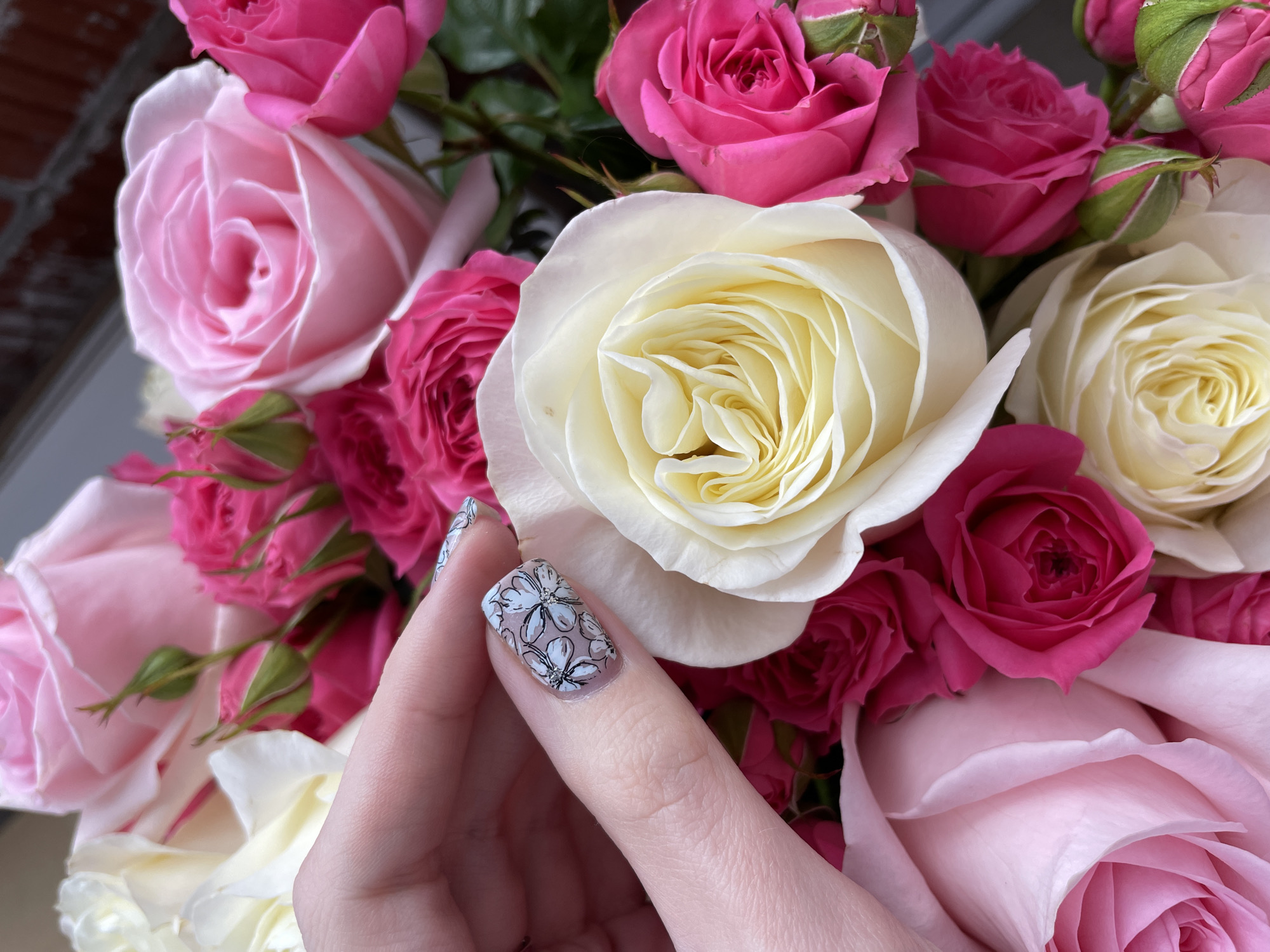 This media file presents a detailed close-up of a vibrant bouquet of roses, likely set indoors in Izhevsk, Russia. The arrangement showcases a rich palette of deep fuchsia, soft pastel pink, and delicate creamy white roses. The flowers are fresh and in various stages of bloom, from tight buds to fully open blossoms, revealing their intricate, layered petals.
In the foreground, a left hand gently interacts with the bouquet, with the index finger and part of the thumb prominently displayed. The index fingernail features an elaborate and artistic nail art design. This design consists of multiple small, light-colored blossoms with intricate black outlines and subtle dark centers, creating a delicate lace-like floral pattern against a translucent or light nude base.
The lighting in the image is bright and natural, suggesting a daytime setting. The background is softly blurred, drawing the viewer's attention directly to the vivid colors of the roses and the detailed nail art. The overall composition evokes a sense of beauty, elegance, and appreciation for both natural aesthetics and personal adornment. The scene captures an intimate moment of admiring or holding the floral arrangement. No visible text is present in the image.