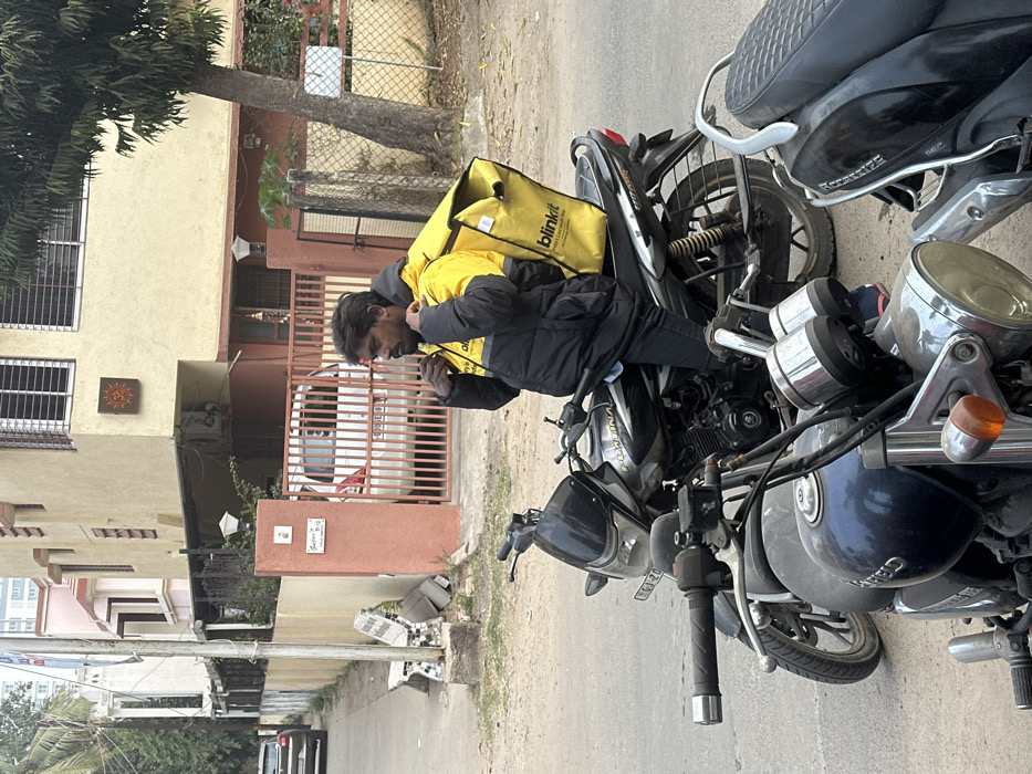 A delivery driver wearing a black and yellow jacket and carrying a "blinkit" delivery bag stands next to his motorcycle. He's looking down, possibly at his phone. The motorcycle is parked on a street, with a building and tree in the background. Another black scooter is parked nearby. The scene takes place during daylight.