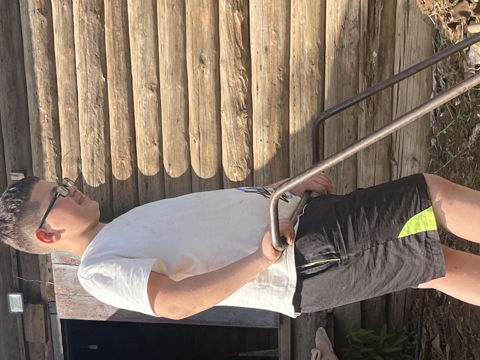 An outdoor, medium shot captures a young Caucasian male, likely in his pre-teens, standing outdoors and operating a gardening tool. He is positioned on the right side of the frame, facing left, with his body angled. He wears a white t-shirt and black shorts with a bright green accent on the right leg. He has short, dark hair and is wearing dark-rimmed glasses. His right hand is gripping the handle of the gardening tool, which is a metal implement with two rods extending from a handle, suggesting it is a rake or a similar hand tool used for gardening. He is looking downwards, focused on his task. Behind him, a rustic, wooden structure dominates the background. The structure appears to be made of horizontally stacked logs, with a doorway or opening visible on the left side of the image, showing some weathered wooden planks. The environment suggests a rural or semi-rural setting, possibly a backyard or garden. The lighting indicates it is daytime, with sunlight casting shadows, particularly on the wooden structure. The overall impression is of a boy engaged in yard work on a sunny day. The location is the city of Фоки, Russia.