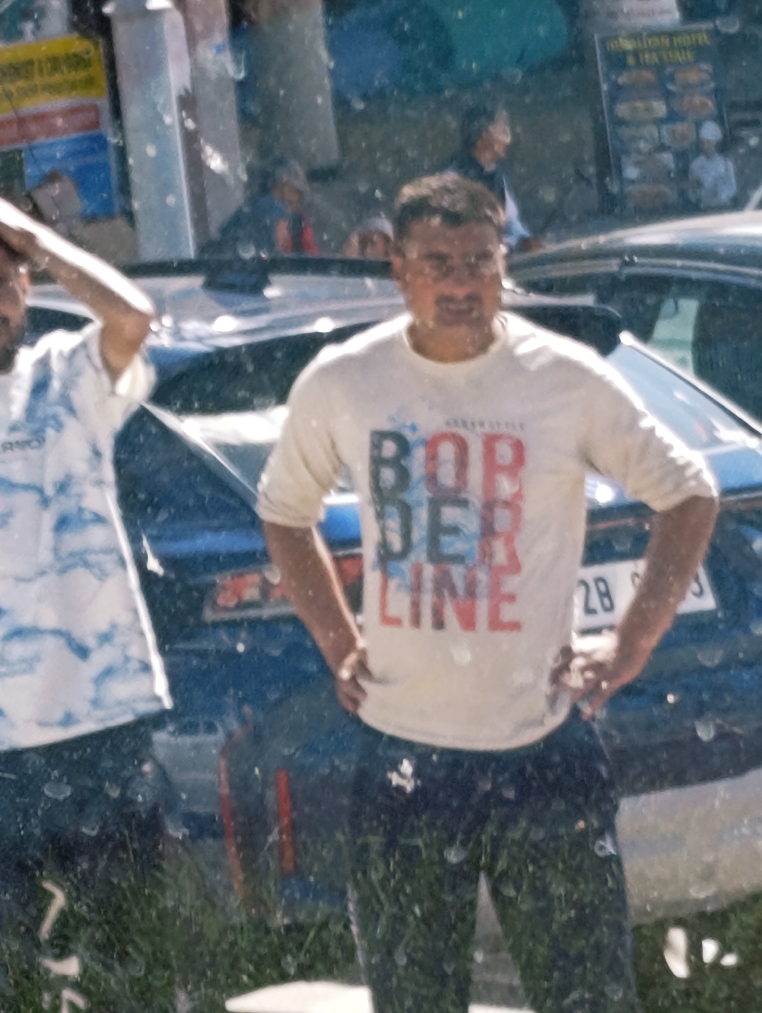 This image captures a reflection of a street scene in Leh, India, likely taken from within a vehicle due to the prominent water droplets on the glass surface distorting the view. The primary focus is on two men. The man in the foreground, wearing a white t-shirt with "BORDER LINE" printed vertically in red and blue, is looking towards the viewer with a slight smile. He has dark hair, a mustache, and is wearing glasses. His right hand is on his hip. To his left, another man is visible, wearing a white t-shirt with a blue and white abstract pattern and dark pants. His left hand is raised, and his face is partially obscured. In the background, reflections of a car with a license plate ending in "2B" and a menu board for "INDIAN HOTEL & RESTAURANT" are discernible. Other blurry figures and elements suggest an urban environment. The presence of a car and the sign point to a location along a road or in a parking area in Leh.