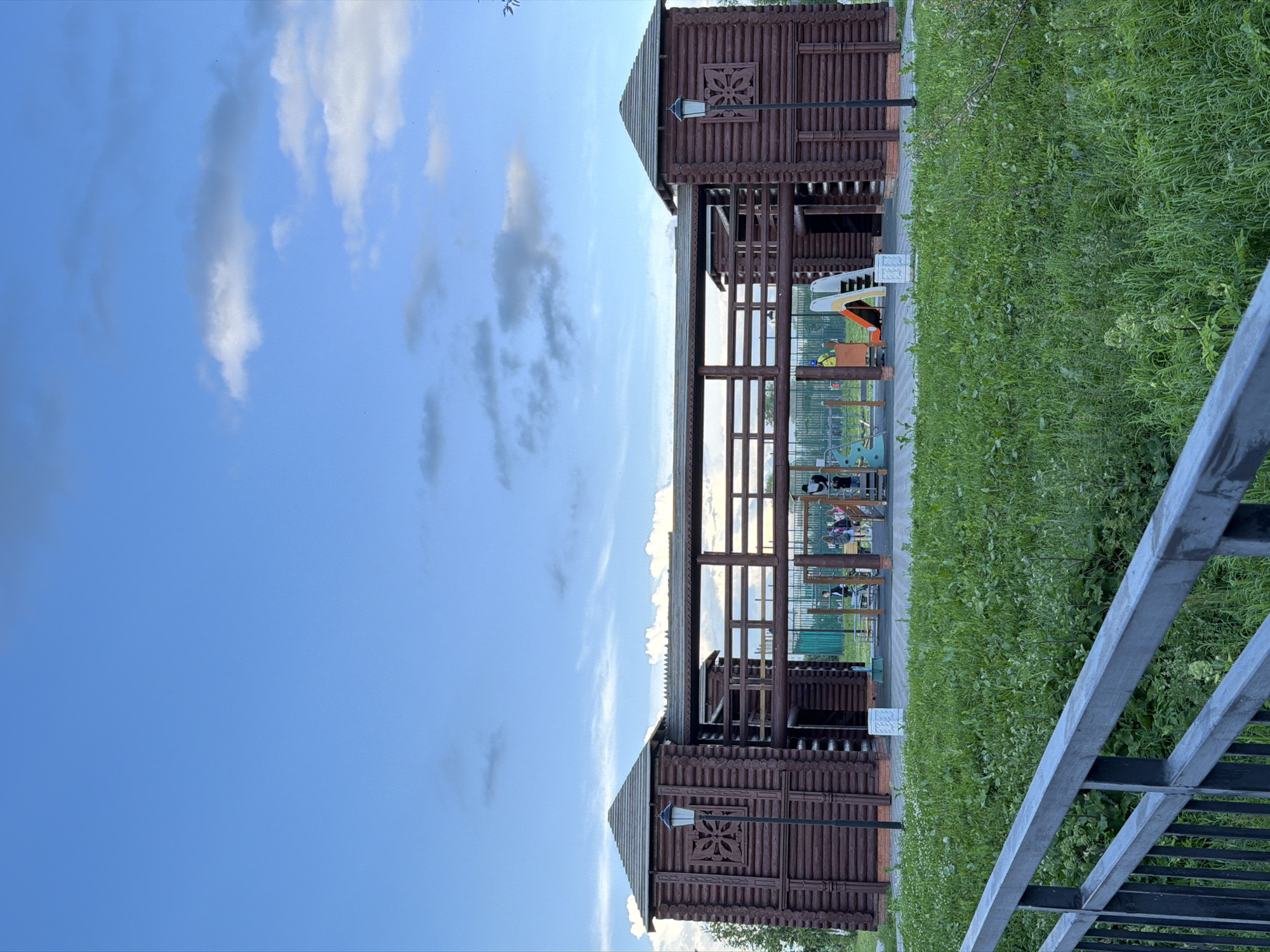 The image shows a playground area with wooden structures reminiscent of medieval fortifications. Two tall, brown, wooden buildings with decorative carvings and pitched roofs stand on either side of a covered walkway. The walkway is supported by wooden beams and has a fence-like railing. Inside the covered area, there is a playground with colorful slides and structures, some of which appear to be climbing apparatuses. Several people, including children and adults, are visible in and around the playground. The ground is paved, and there is a lush green area with tall grass adjacent to the playground. A metal fence runs along the right side of the image, partially obscuring the view. The sky is a bright blue with scattered white clouds, suggesting it is daytime and the weather is fair. The overall setting appears to be a park or recreational area in Solikamsk, Russia. There is no visible text in the image.