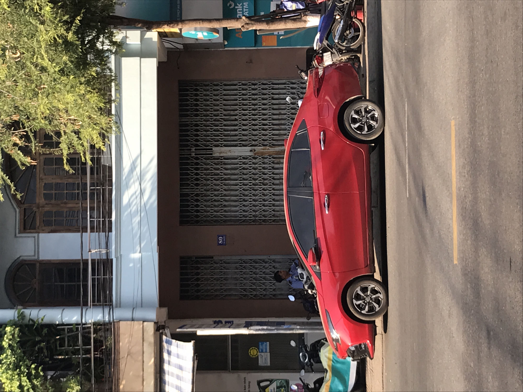 A red car is parked on the street in front of a building in Nha Trang, Vietnam. The building has a gray metal roll-up door, windows, and a sign with the number "80". Several parked motorbikes are in front of the building, and a few people can be seen in the background. A tree with green foliage is present on the left side of the frame. The street is paved. A yellow line is visible on the side of the road.