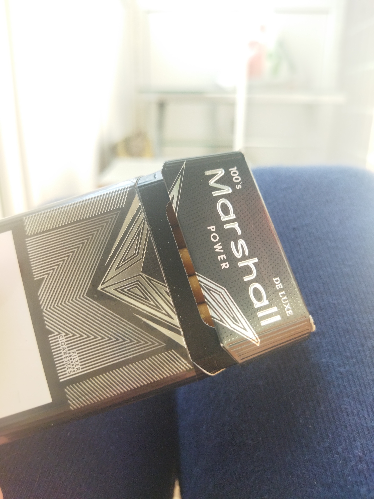 A pack of "Marshall" brand cigarettes, specifically "100's Power De Luxe", is partially open, revealing the ends of the cigarettes inside. The pack is black with silver geometric designs and lettering. It rests on a surface covered with blue fabric. No people are visible. There is no indication of the location.