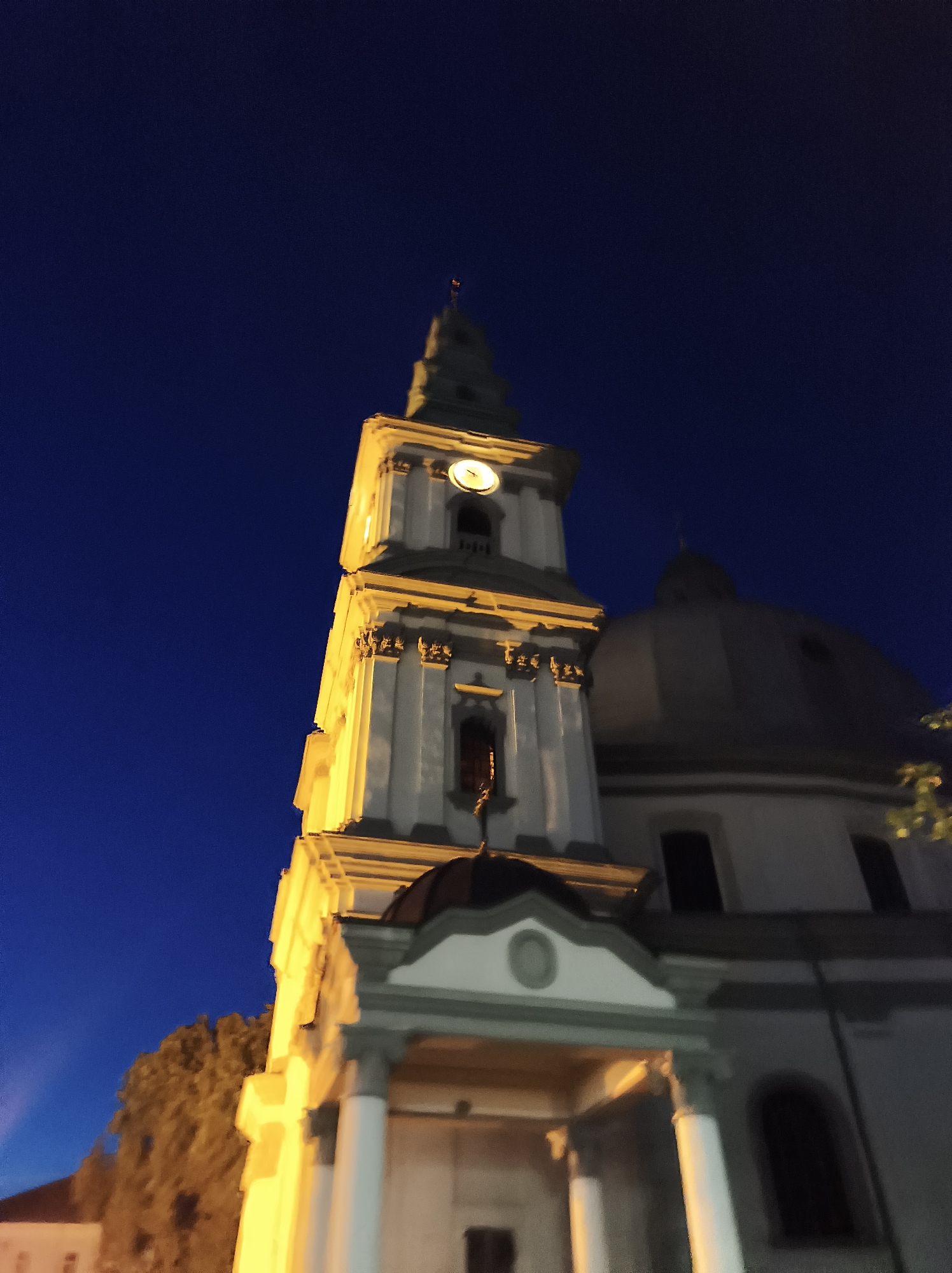 The image depicts the Ternopil Theological Seminary's bell tower and dome at night. The architecture features a tall, multi-tiered bell tower with a clock face illuminated on one side. It is adorned with classical columns and ornamental detailing, all bathed in warm artificial light. Adjacent to the tower, a large, dark dome rises against the deep blue night sky. The lower part of the building reveals a portico with prominent white columns and arched windows. To the left, a silhouette of a tree with foliage is visible, with the faint outline of another building in the background. The overall scene is dimly lit, with the primary illumination coming from the building's own lighting.