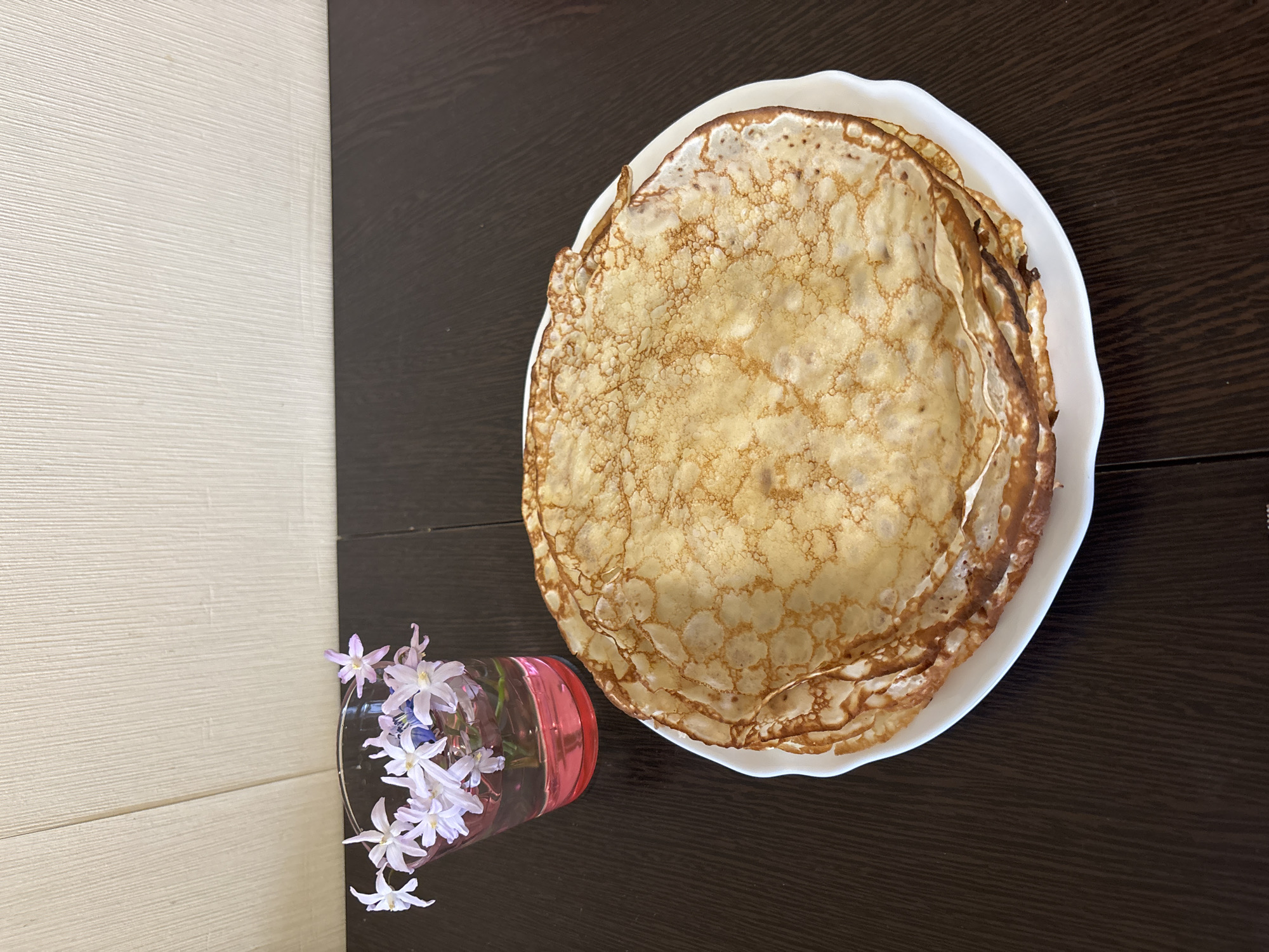 The image presents a top-down view of a still life arrangement. On the right, a stack of crepes sits on a white plate. To the left, a vase of small, light-purple flowers rests on the surface. The background is a split surface, with a light-colored, textured wall on the left and a dark-brown wooden table on the right. Based on the location information, this scene is presumed to be in Chaikovsky, Russia.
