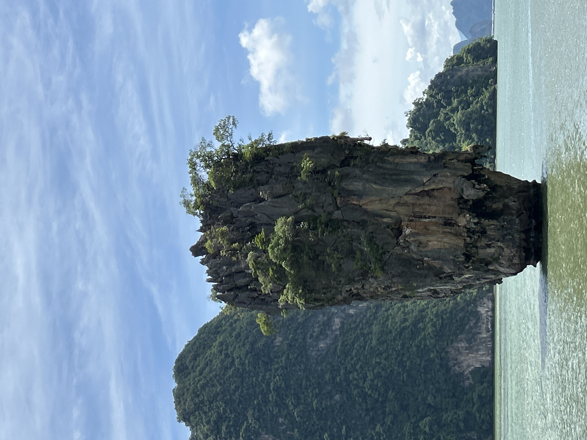 The image shows a tall, narrow limestone karst island in the middle of the sea, a characteristic feature of Phang Nga Bay in Krasom, Thailand. The island is covered in lush green vegetation, with trees growing precariously on its rocky surface. The water around the island is a tranquil turquoise-green, with gentle ripples reflecting the blue sky and scattered white clouds. In the background, other similar karst islands and lush green hills rise from the sea, creating a dramatic and picturesque landscape. The scene is bathed in natural sunlight, suggesting a clear or partly cloudy day. There are no people or boats visible in the immediate vicinity of the main island. The overall impression is one of natural beauty and geological wonder.