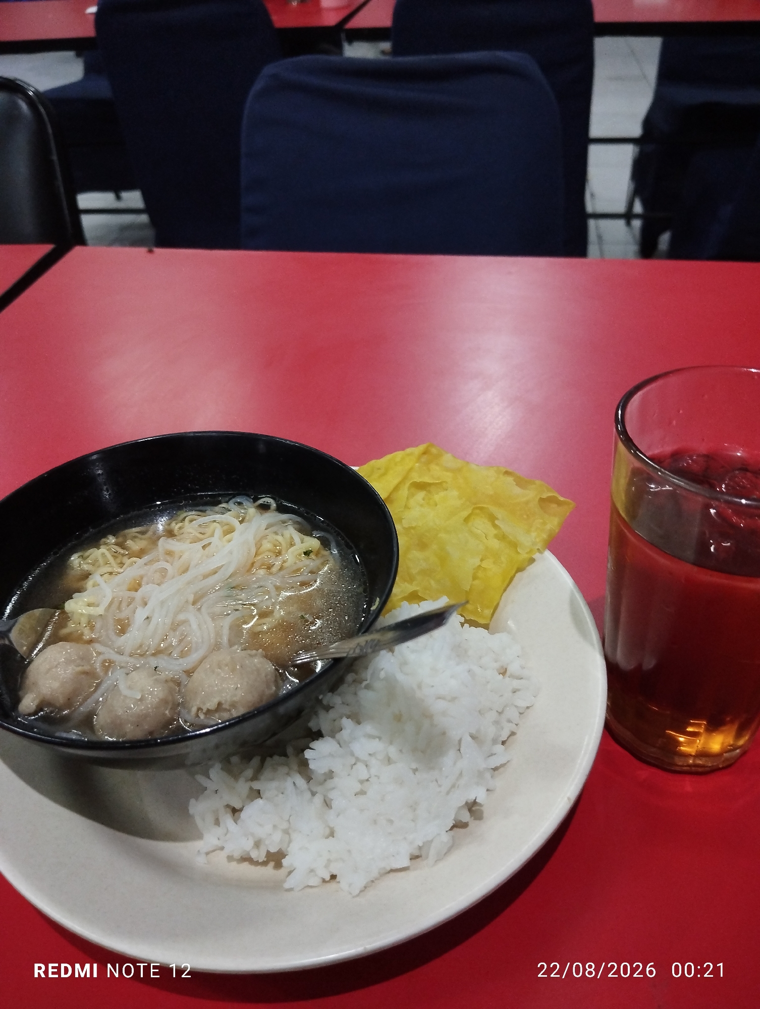 A plate of food and a glass of iced tea are on a red table in a dimly lit room. The plate contains white rice, a bowl of noodles with meatballs, and a fried wonton. The glass of iced tea is on the right side of the plate, and the room appears to be a cafe or restaurant. The timestamp on the image indicates it was taken on August 22, 2026, at 00:21. The text at the bottom left of the image "REDMI NOTE 12" likely indicates the phone used to take the photo. The background shows out-of-focus dark blue chairs, suggesting a seating area. The overall impression is a simple meal set on a table in a public eating establishment.