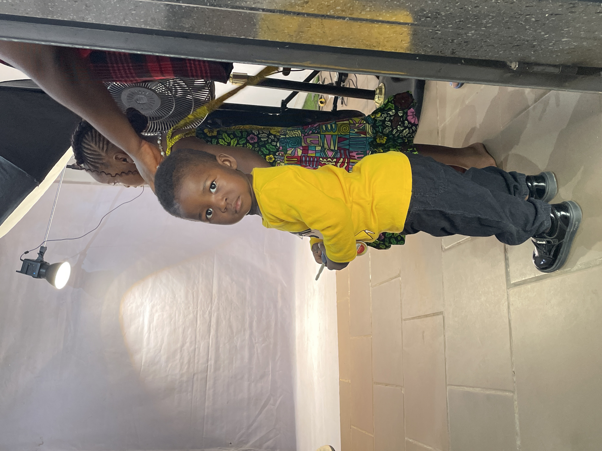 A medium indoor shot depicts a child in a yellow shirt and dark pants, standing with an adult woman wearing a colorful patterned dress. The child is looking at the camera. Behind them is a white backdrop illuminated by a spotlight. A fan is visible in the background. The location is likely a studio, possibly in the city of Jos, Nigeria.