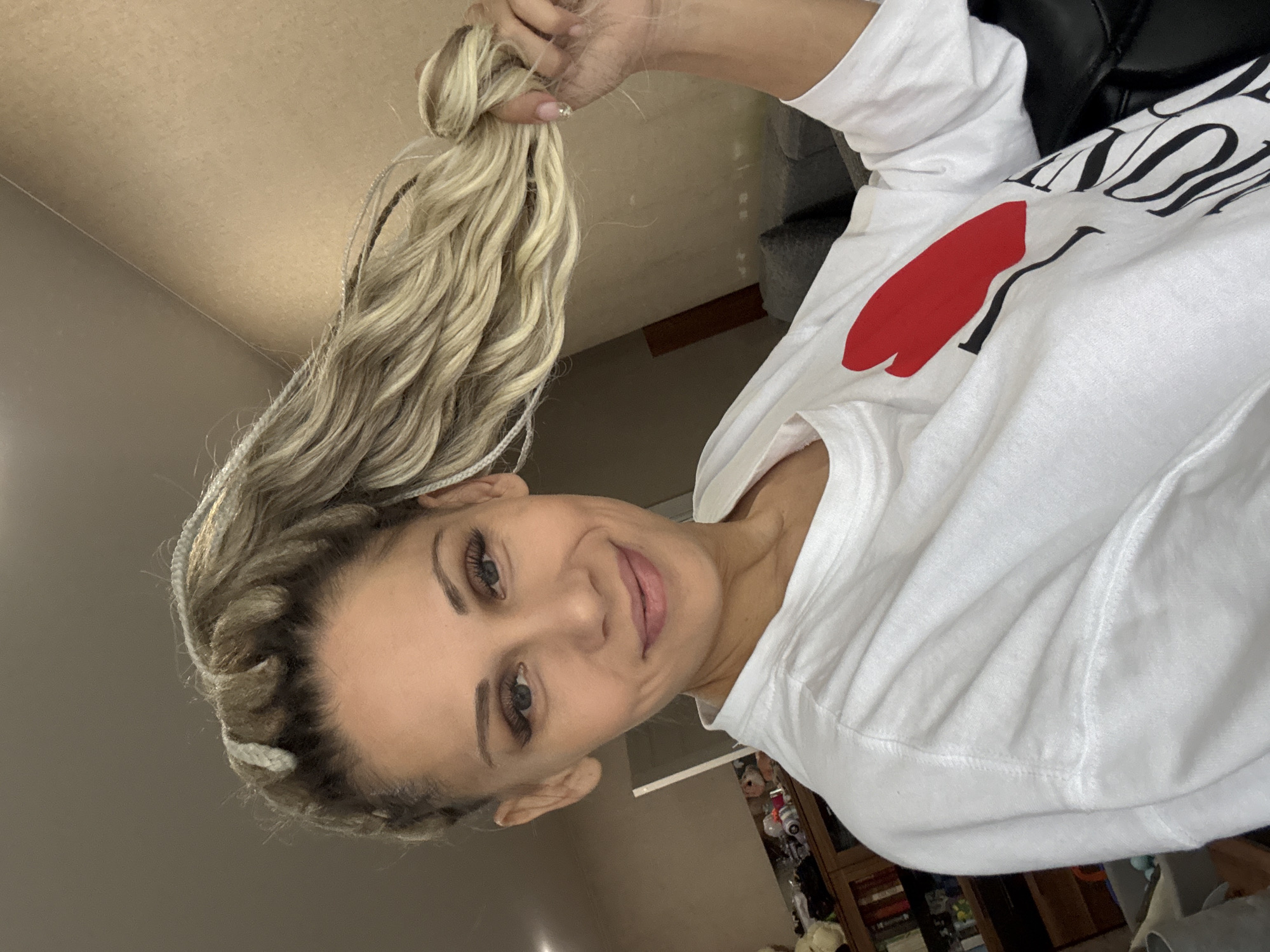 A woman with blonde braided hair is holding her hair up in her right hand. She is wearing a white sweatshirt with a red heart and black lettering. The background is a beige wall. The location, Rusanovo, Russia is not discernible from the visual information in this image.