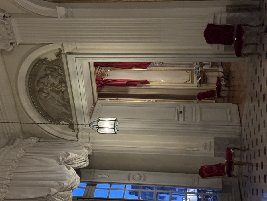 A room with elaborate architectural details and decorations. The ceiling has ornate molding and carvings, including a semicircular relief of figures. A chandelier hangs from the ceiling. Red curtains are visible in a doorway, and several antique chairs are placed in different areas of the room. Windows are at the bottom, showing a cityscape with blue sky. The floor is tiled with a checkered pattern.