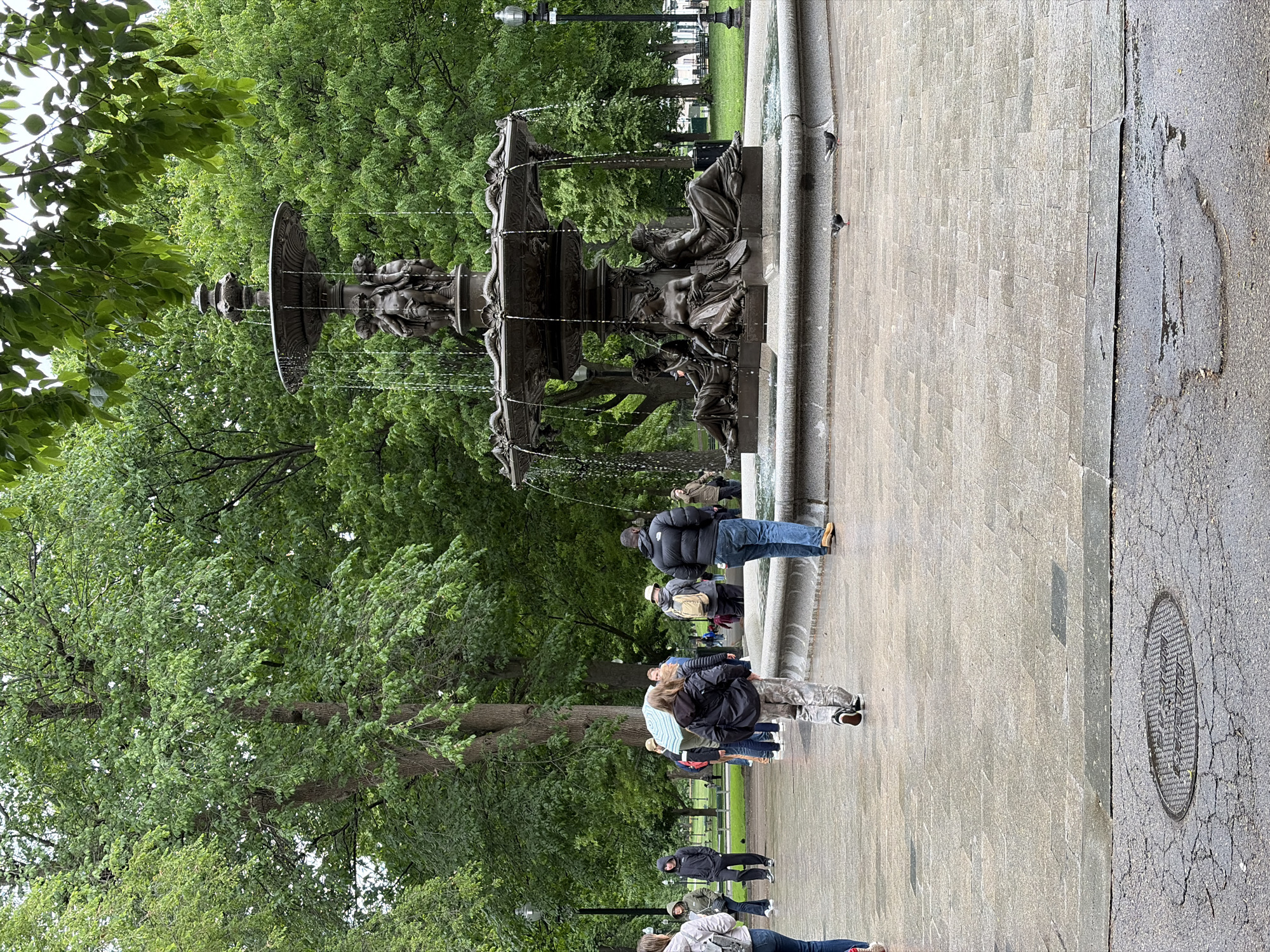 The image depicts the Brewer Fountain, located in Boston Common, Boston, Massachusetts. The fountain is made of dark metal and features a multi-tiered design with sculptures and water flowing from each level. A group of people are standing near the fountain, seemingly observing or taking pictures. The scene is set on a paved area adjacent to the fountain, with trees and green foliage visible in the background. A sewer cover is also visible to the right of the fountain. The overall atmosphere appears to be a typical day in a public park.