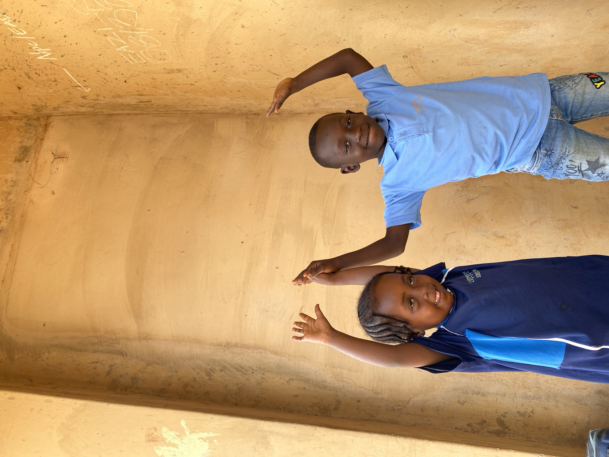 Two children, a boy and a girl, are positioned facing the camera indoors in Kuru Dungus, Nigeria. The background consists of yellowish-beige walls. The boy is in the upper right, wearing a light blue collared shirt and patterned jeans. The girl is in the lower right, wearing a navy blue top. Both children have their arms raised with hands open. The image is rotated to the left. There is writing on the wall behind the boy. The walls appear to be weathered.