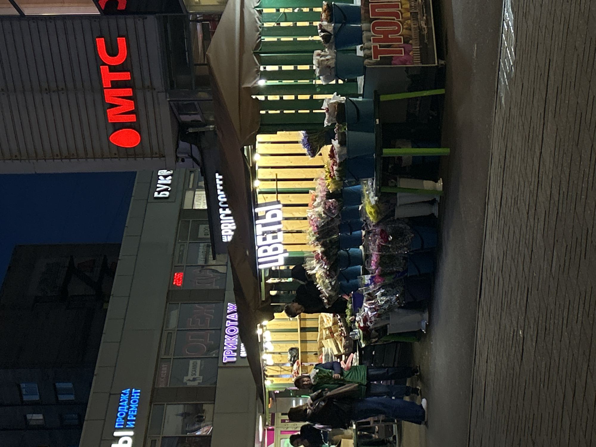 The image depicts an evening street scene in Saint Petersburg, Russia. A flower stall, featuring bouquets in buckets, stands on the sidewalk. Shoppers are browsing and interacting with the merchandise. Storefronts with signage in Russian, including "МТС" (a mobile network operator) and "ЦВЕТЫ" (flowers), line the street. The brick pavement suggests an urban environment.