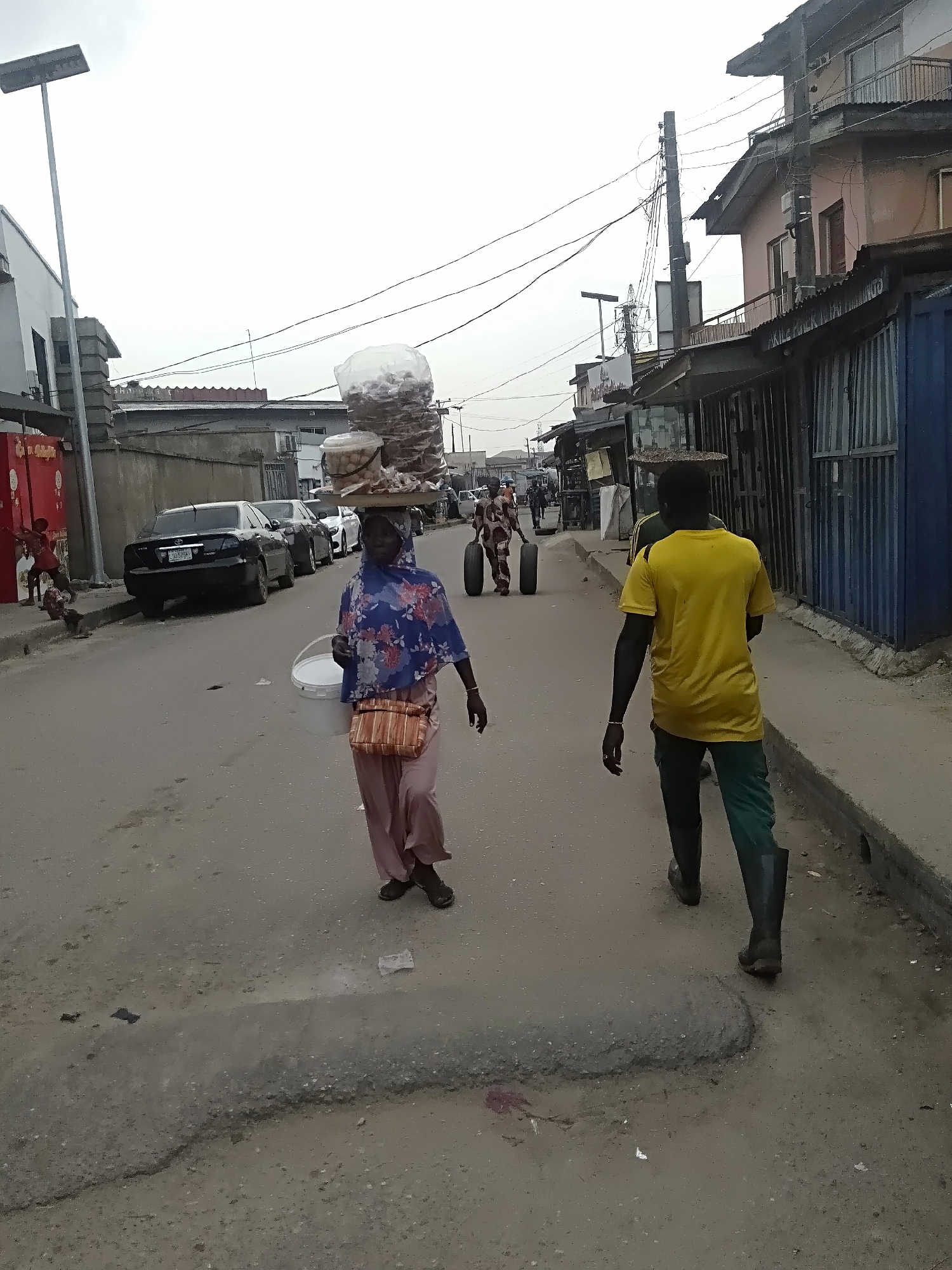 This image depicts a bustling street scene in Shangisha, Nigeria, under an overcast sky during the daytime.
In the foreground, a woman walks towards the left side of the frame, expertly balancing a large wooden tray laden with various packaged goods, including snacks and what appear to be nuts in clear plastic bags and containers, on her head. She wears a blue floral head covering, a blue top, and light pink pants, and carries a white bucket in her left hand and a striped woven bag slung over her right shoulder. To her right, a man in a bright yellow t-shirt, dark green pants, and black boots walks away from the camera.
The street is paved but shows signs of wear, with visible cracks and a rough, raised section of concrete in the immediate foreground. Cars are parked along the left side of the street, including a black sedan with a partially visible license plate starting with "GDD-5...". Buildings line both sides of the road, featuring a mix of commercial establishments and residences, characterized by corrugated metal roofs, some balconies, and a dense network of overhead utility wires. Further down the street, another person is seen transporting two large tires, and children are visible near a red structure on the left. A visible sign on a blue corrugated metal structure on the right side of the street appears to read "AKILLE POWER TAI... ENTERPRISE". The scene captures typical daily activity and commerce in an urban Nigerian setting.