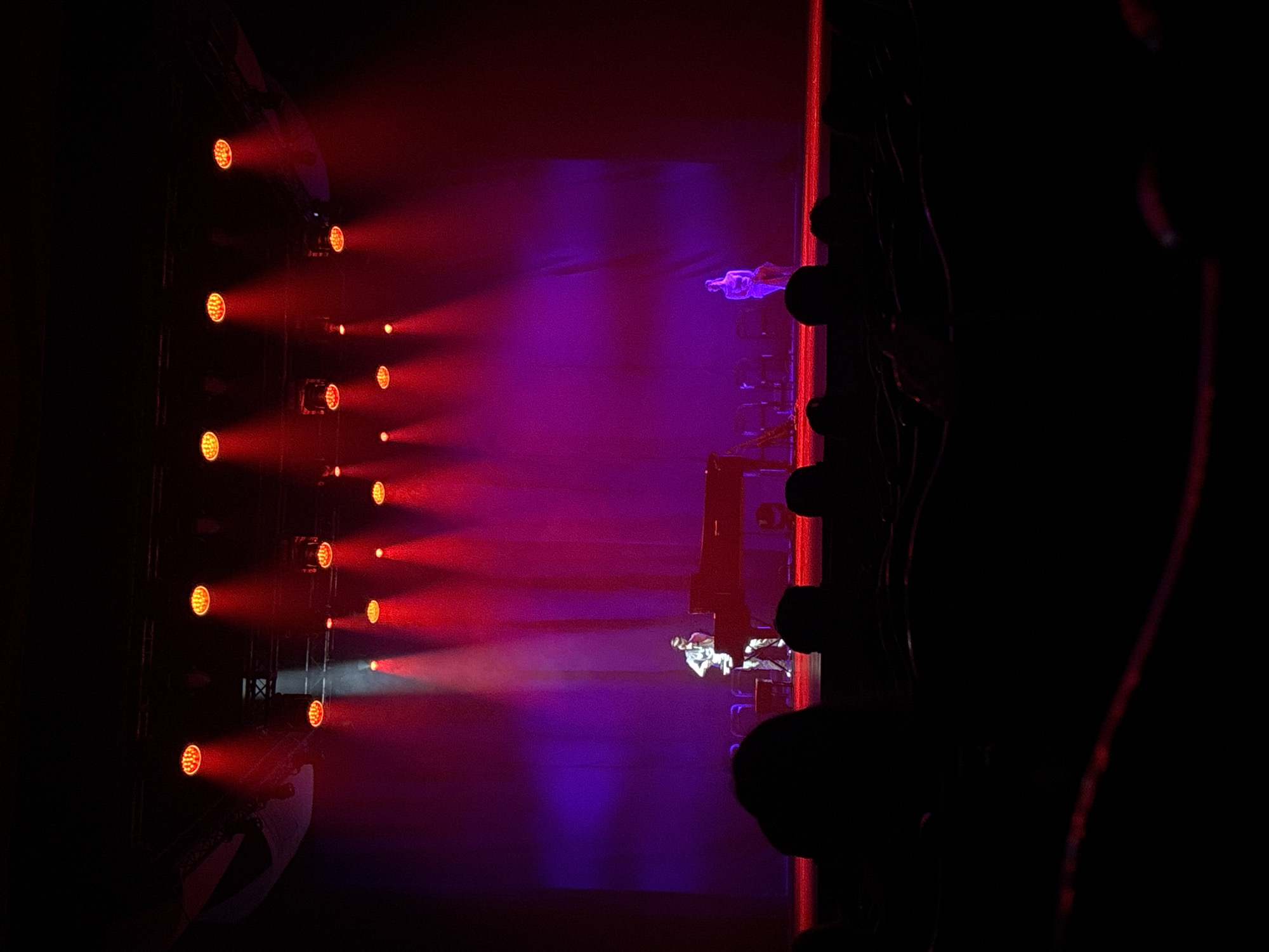 The image shows a theatrical stage in Perm, Russia, lit with a combination of red and purple lighting. A grid of round, red stage lights are visible on the left, with their beams casting a red hue across the stage. Two figures, dressed in white attire, are positioned on the stage: one near the foreground and another farther back. Silhouetted elements of the theater's infrastructure are present on the right side of the frame, adding depth and obscuring details in that area. The overall ambiance is dark and dramatic.