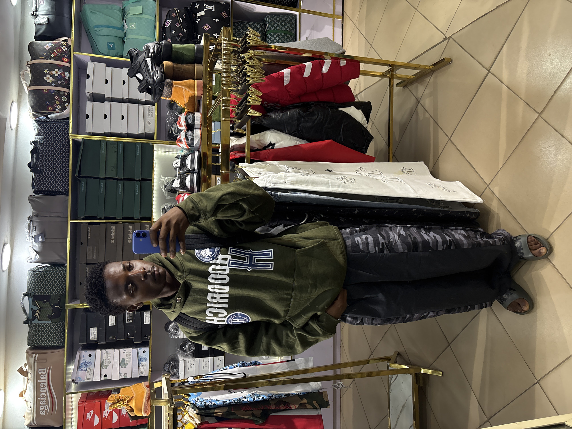 A young man takes a selfie in a retail store in Bukuru, Nigeria, capturing his reflection in a blue smartphone. He is wearing an olive green hoodie with "HOODRICH" and "DIMIT MON" text and a circular logo, camouflage-patterned pants, and dark slide sandals. The store features multiple shelves stocked with various merchandise, including clothing, bags, and shoes.
To the left, several bags are displayed, including a beige Balenciaga duffel bag, a bag with an LV monogram pattern, and another with a green Goyard-like pattern. Above these, there are teal and black backpacks. On the right, gold-colored clothes racks hold stacked jackets in red, black, and white, as well as folded shirts. The shelves also contain numerous shoe boxes, some clearly marked with "NIKE" and "Timberland" logos. Other footwear includes various sneakers, black sandals, and tan boots. The floor is tiled with light-colored square tiles laid in a diagonal pattern. The store is brightly lit by overhead lights, one of which is visible on the left side of the frame. The man's expression is neutral as he looks at his phone.