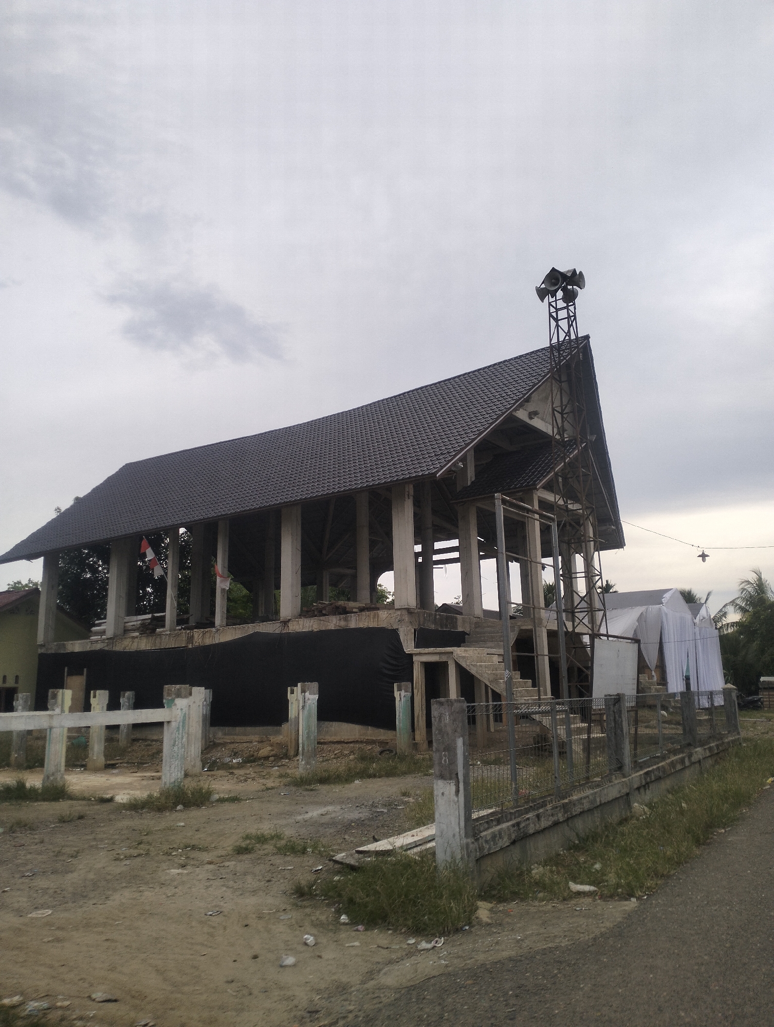 The image depicts an unfinished building with a dark grey tiled roof and visible concrete pillars. A metal tower with multiple speakers is mounted on the roof's peak. Two Indonesian flags are displayed on the second level of the structure. In the foreground, a dirt path is visible with patches of dry grass, and a low concrete barrier with a wire mesh fence runs along the side. To the right, a road is present. Behind the main structure, there are white tent-like coverings and palm trees. The sky is overcast with grey clouds. The scene appears to be an outdoor location in a semi-urban or rural setting under construction.