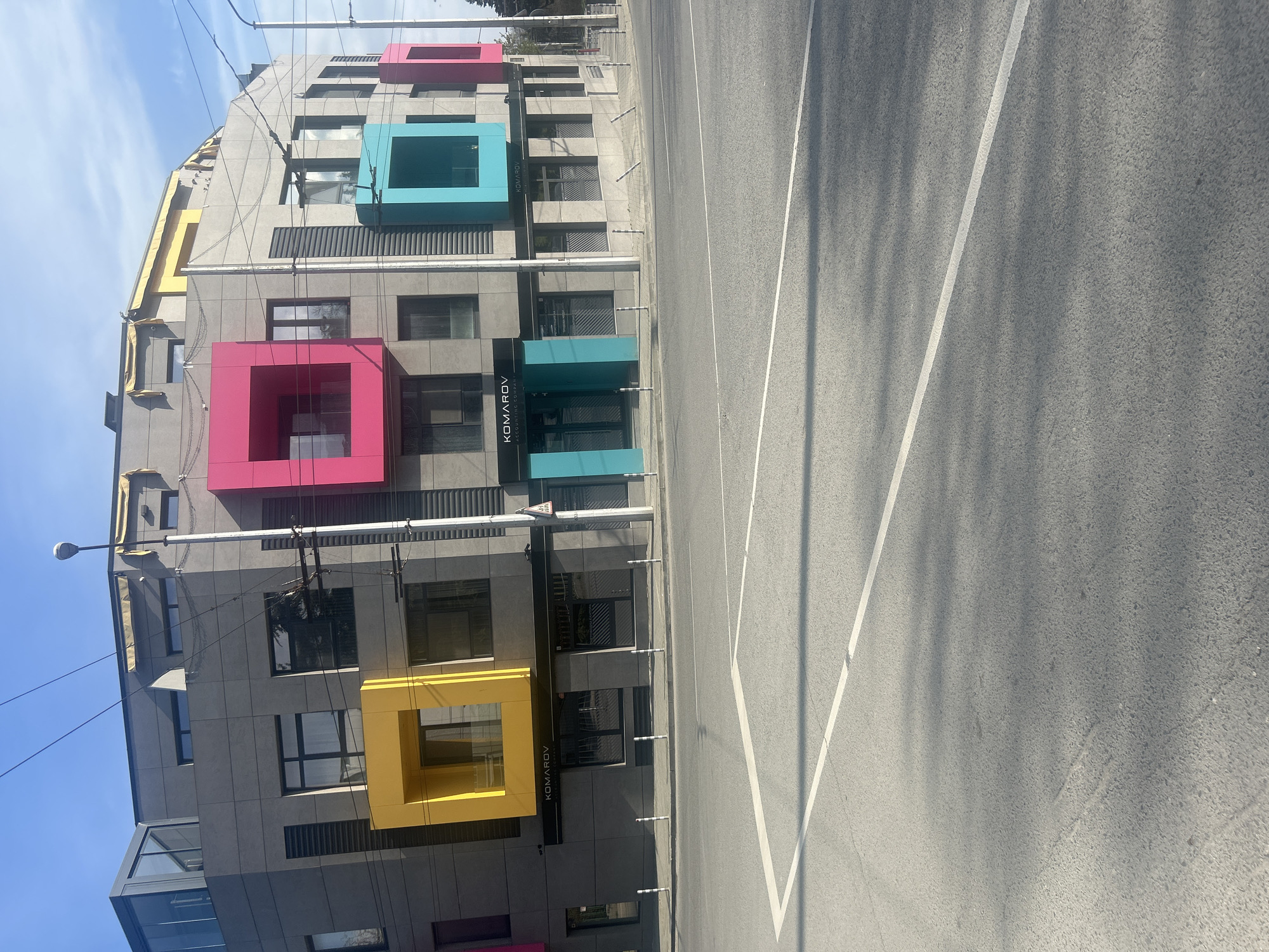 The image depicts a multi-story building with a modern architectural design, characterized by a gray facade punctuated by colorful square projections. These squares are painted in shades of yellow, pink, and blue. The building appears to be located on a city street, evidenced by the presence of a marked road with white lane lines. The name "KOMAROV" is vertically displayed on the building's exterior. The building is located in Varna, Bulgaria. Overhead power lines are visible above the building. The sky is partially clear with some cloud cover. The scene is bathed in daylight, with shadows cast on the road surface.
