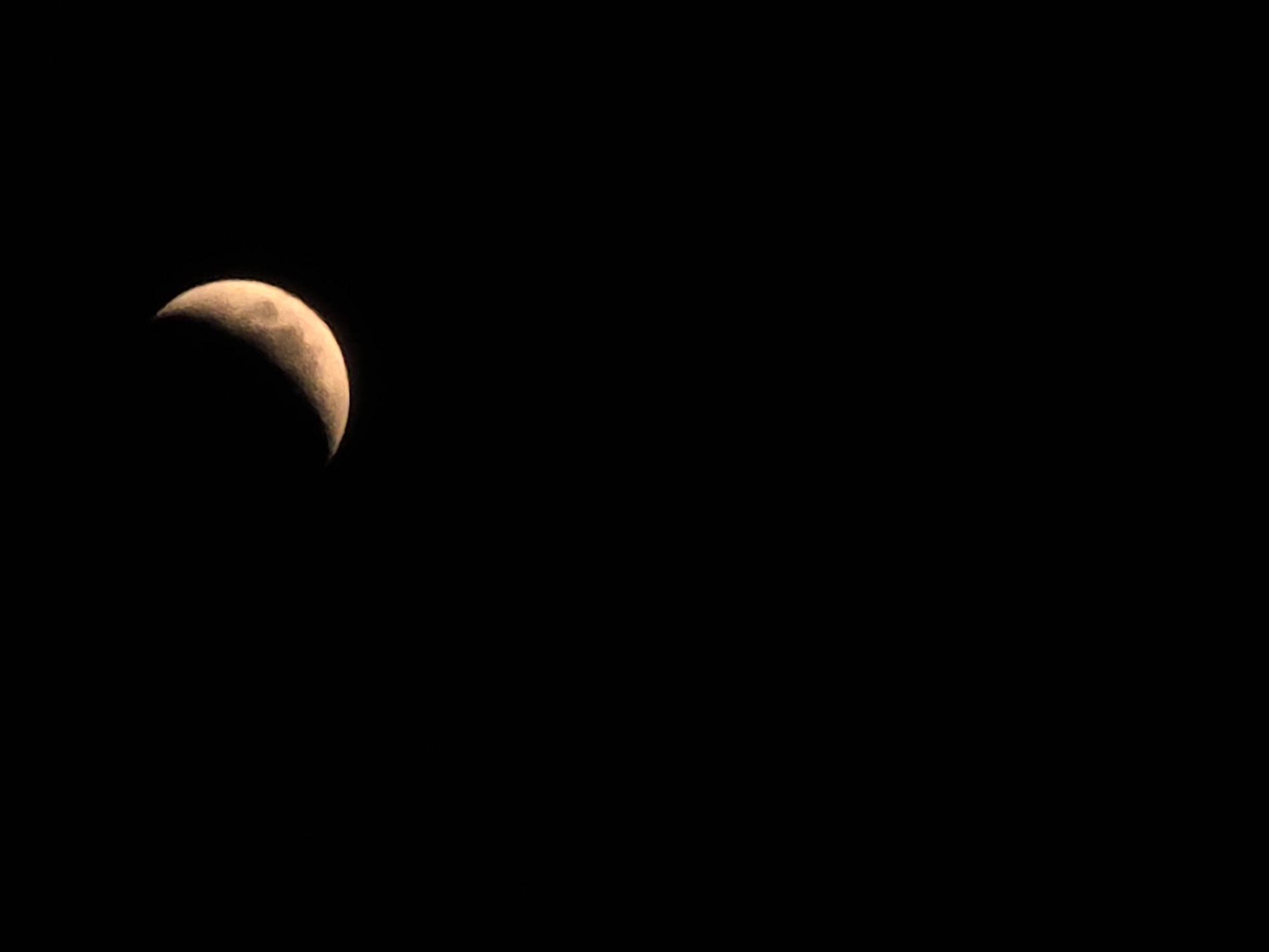 A crescent moon is visible in a dark sky. The moon is partially illuminated, with a bright yellow-orange glow on its left side and a shadowed, darker crescent on its right. The sky is completely black, indicating it is nighttime. The moon is positioned in the upper right quadrant of the image.