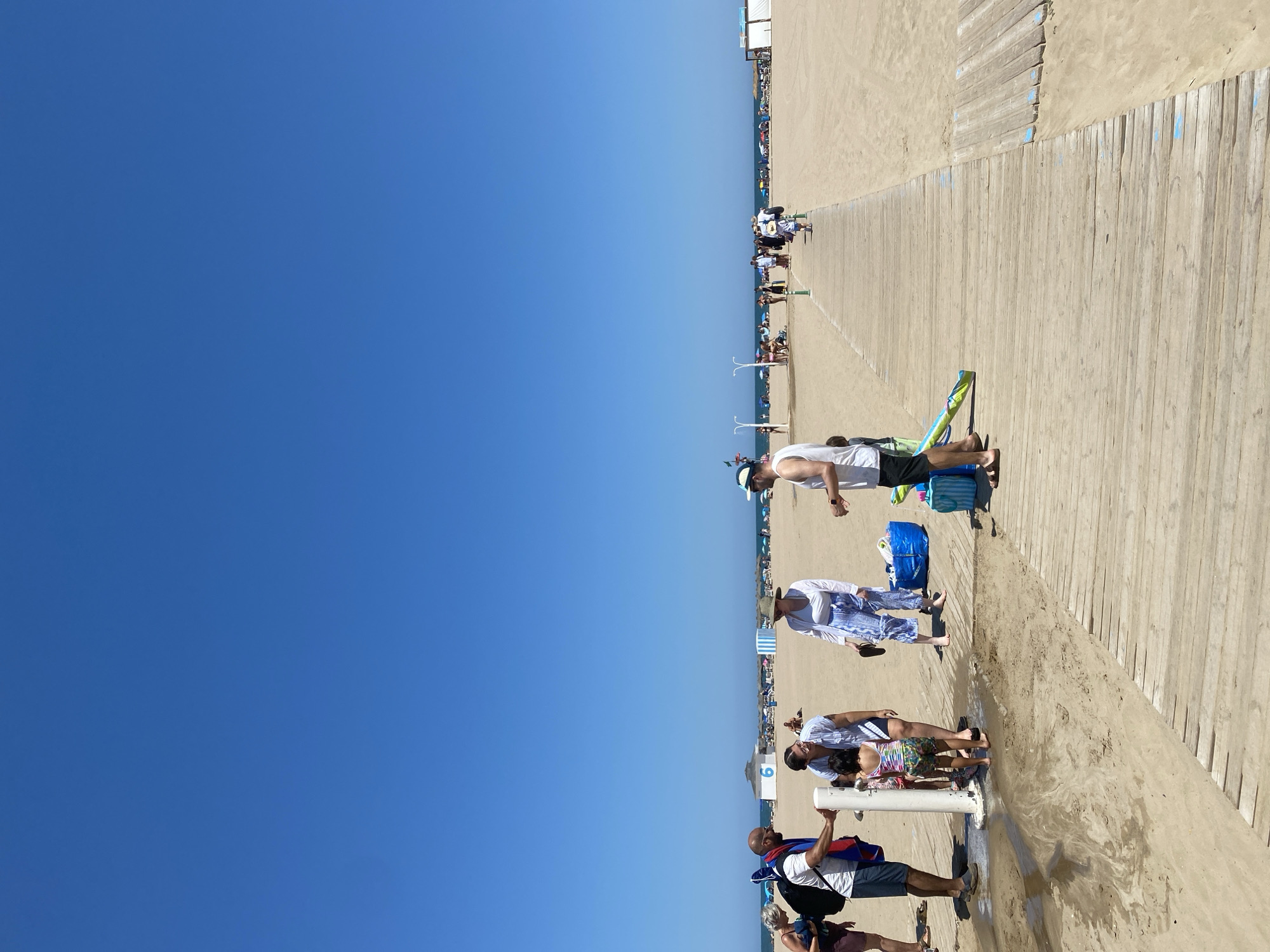 The image shows a wide view of a sunny beach in Valencia, Spain. The scene is dominated by a clear blue sky and a sandy beach with a wooden boardwalk. Several people are visible on the boardwalk and on the sand, some walking, others appearing to be relaxing.
In the foreground, a person in a white tank top and black shorts is leaning over, possibly adjusting something on the boardwalk. Further down the boardwalk, a group of people are gathered. One person, wearing a white shirt with blue patterns and a hat, stands next to another individual in a similar white outfit. They appear to be interacting. Closer to the water's edge, a group of people are around a public shower or faucet, with one person holding onto it.
In the background, there are many beach umbrellas and loungers, indicating a busy day at the beach. The ocean is visible in the distance, stretching to the horizon. The weather appears to be hot and sunny, typical for a summer day in Spain. The atmosphere seems relaxed and casual, consistent with a beach outing. There is a number "9" visible on a small structure near the water.