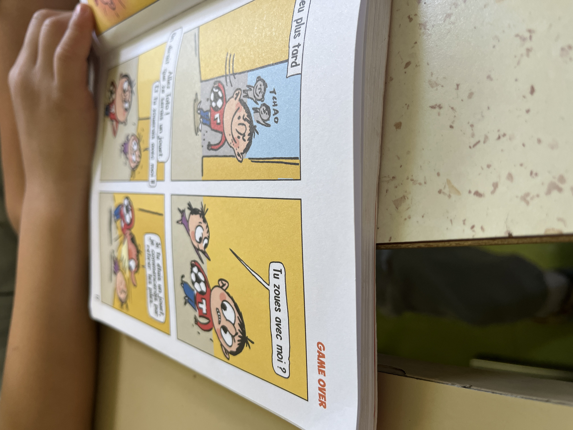 A child's arm and hand are visible on the left, holding open a comic book that rests on a light-colored, speckled table surface. The scene takes place indoors, likely in La Fouillouse, France, given the provided context. The comic book is displayed vertically, featuring several panels with cartoon characters.
In the upper section, a banner reads "eu plus tard" (later). The top-right panel shows a character, wearing a red shirt with a number 7 and a soccer ball, waving goodbye ("TCHAO") to two smaller figures. In the top-left panel, another character speaks a dialogue bubble that reads, "Allez Toto! On dirait que tu serais un jouet" (Go Toto! One would say you'd be a toy).
The bottom-right panel depicts the character in the red shirt looking up at another figure, asking, "Tu zoues avec moi ?" (Do you play with me?). The bottom-left panel features a character saying, "Si tu étais un jouet, je commencerais par retirer tes piles" (If you were a toy, I would start by removing your batteries), suggesting a playful or teasing interaction. The bottom-right corner of the page also prominently displays the text "GAME OVER" vertically.
The table surface shows small, scattered brown speckles, possibly from its design or general use. The background beyond the table is dark and blurry, offering no additional contextual details. The child's activity is simply reading or looking at the comic book.