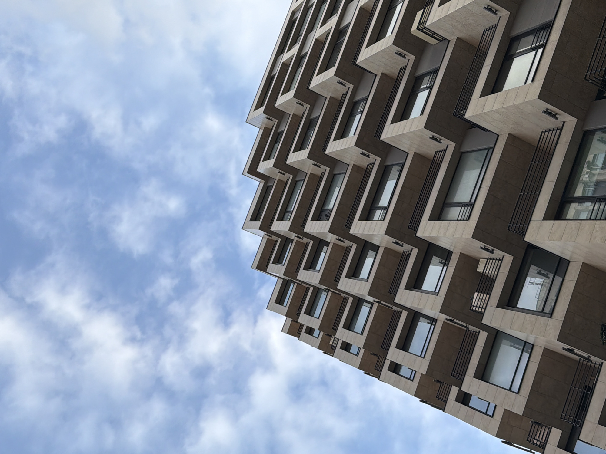 The image shows a modern building in Moscow, Russia, angled upwards against a cloudy blue sky. The building's facade features a unique, staggered design with protruding sections that create a zig-zag pattern. Windows are integrated into each section, and dark metal railings are attached to the exterior. The building's material appears to be a light beige or stone-like composite with darker brown accents. The sky is partially covered with white clouds.