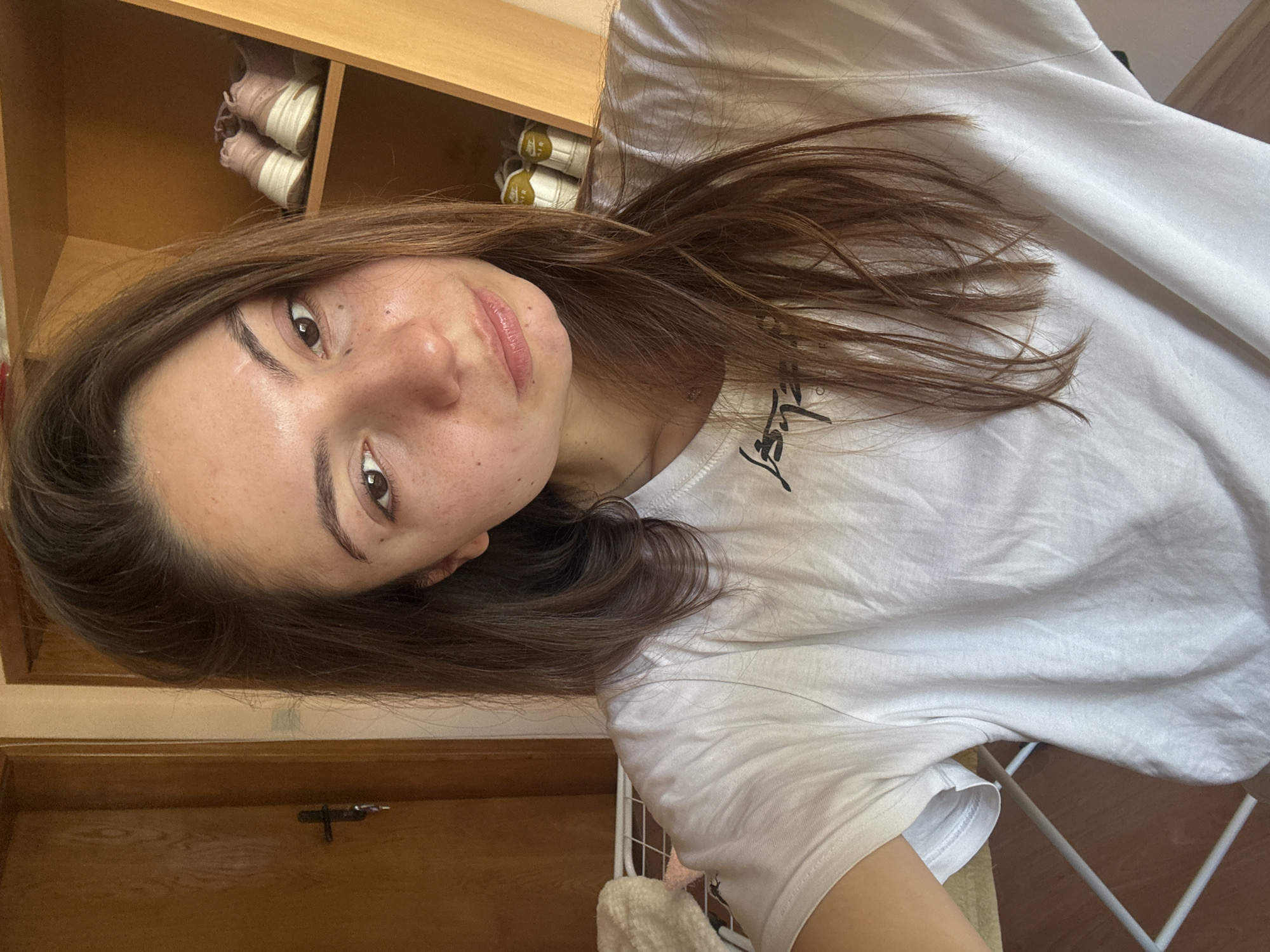 A young woman with long brown hair and freckles on her face is taking a selfie. She is wearing a white t-shirt with a black graphic design. Behind her, there is a wooden closet with shelves containing pairs of sneakers. To her left, a wooden door with a handle and a key is visible. The scene appears to be indoors, likely in a residential setting. There is no discernible information within the image that would allow for identification of the specific location within the city of Varna, Bulgaria.