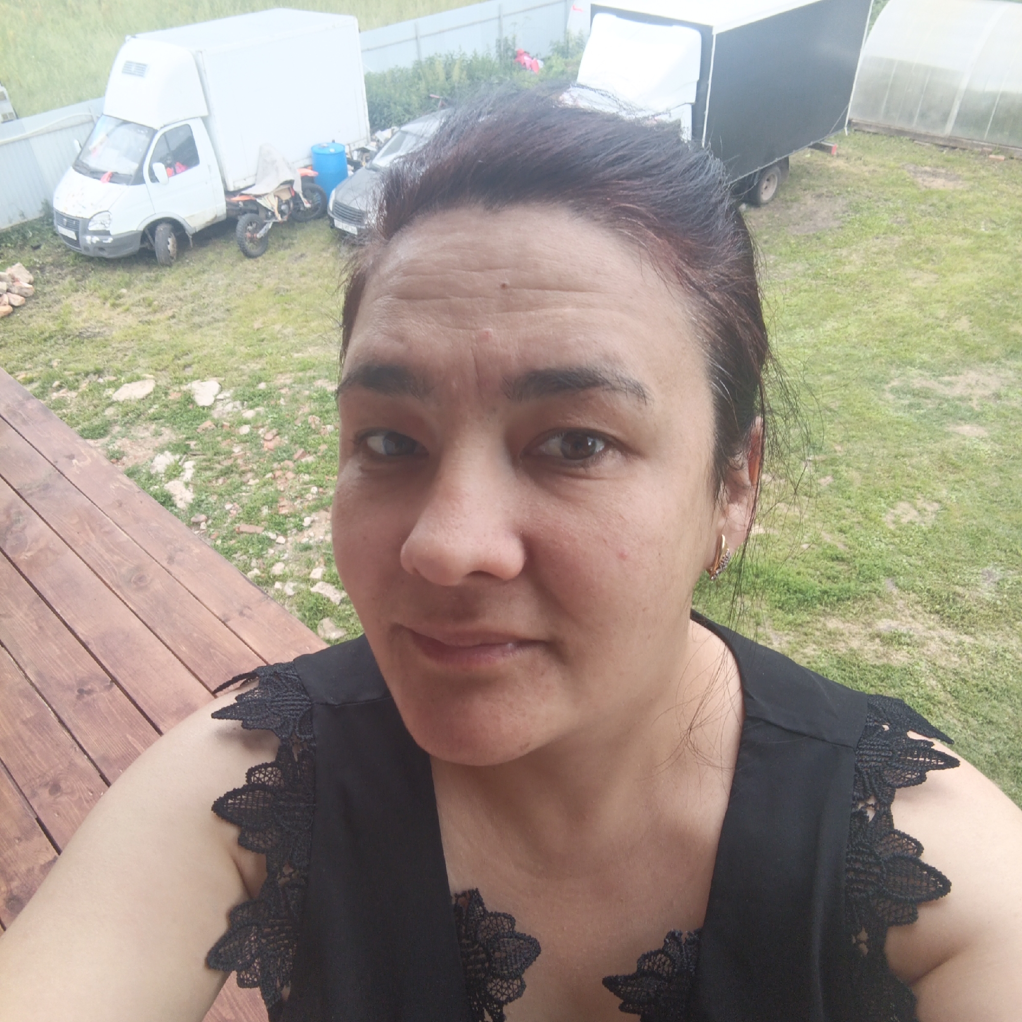 The image shows a woman taking a selfie outdoors, with a wooden deck in the foreground and a grassy yard behind her. In the background, there are several vehicles, including a white delivery truck, a motorcycle, and a black trailer. A greenhouse is also visible. The woman is wearing a black top with lace detailing on the shoulders. She has dark hair and is looking directly at the camera with a neutral expression. The scene appears to be set during the daytime, with overcast skies. The overall impression is of a casual outdoor setting, possibly a rural or suburban area. The location context is identified as Podlubno, Russia. There is no visible text in the image.