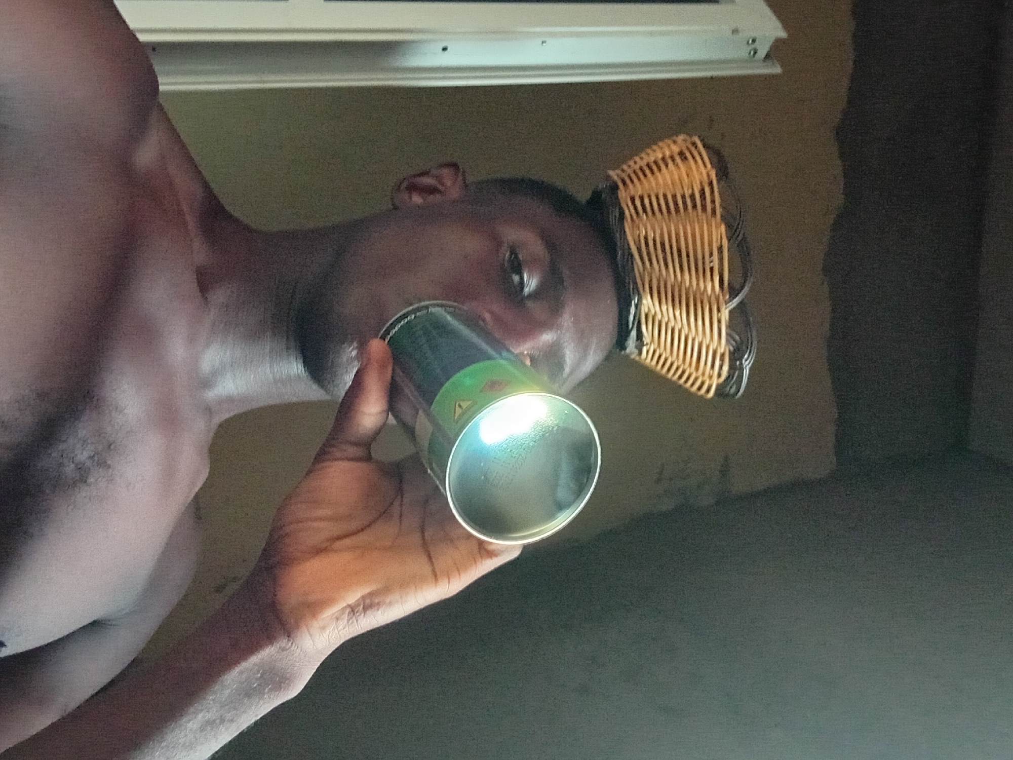 A shirtless man in Akwete, Nigeria is drinking from a silver can. The can has some green and red details on it. He is wearing a woven hat or basket-like object on his head. The background appears to be a wall of a room with a possible light fixture above.
